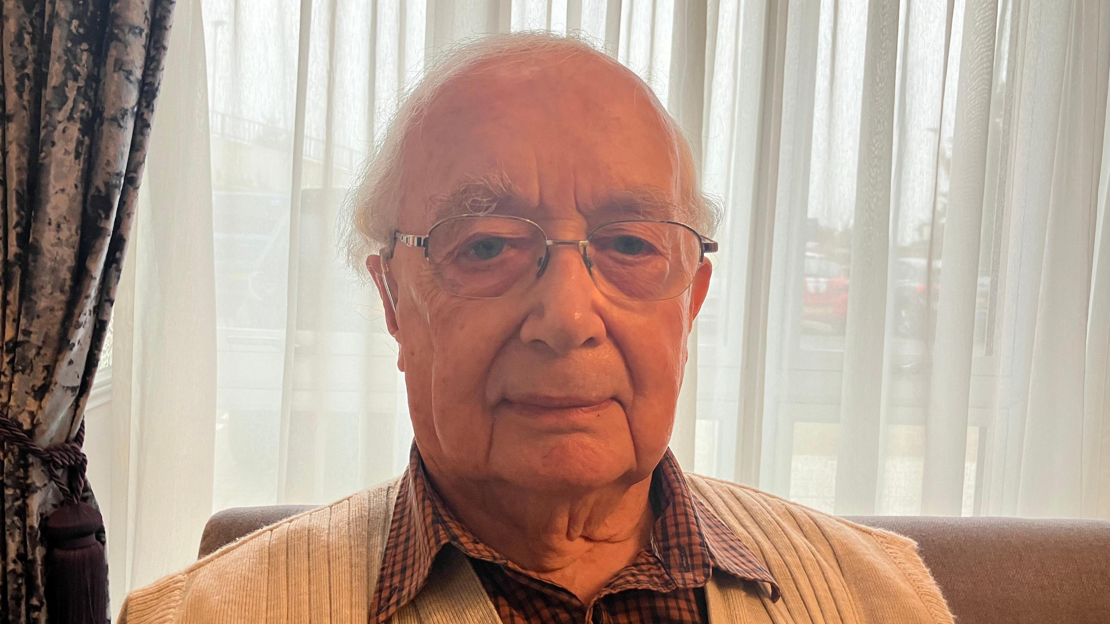 A head and shoulders photo of a man with white hair and glasses, wearing a red checked shirt and a grey cardigan, looking straight at the camera. He's sitting on a sofa and there's a window behind him.