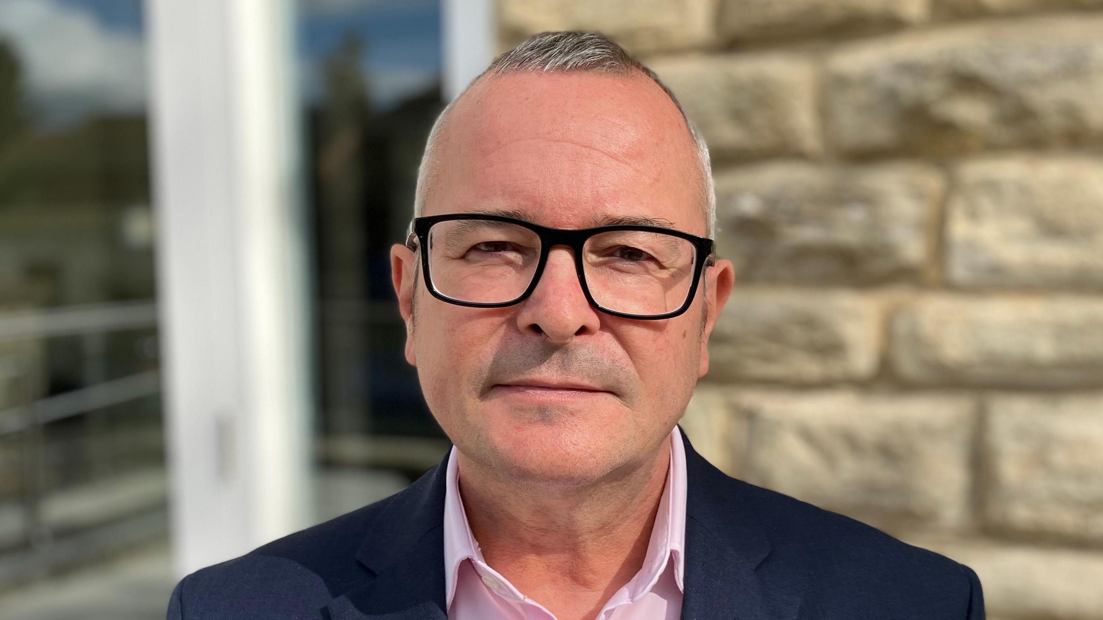 Lee Barron with short white hair and black-framed glasses, wearing a blue jacket and pink shirt standing in front of a stone wall