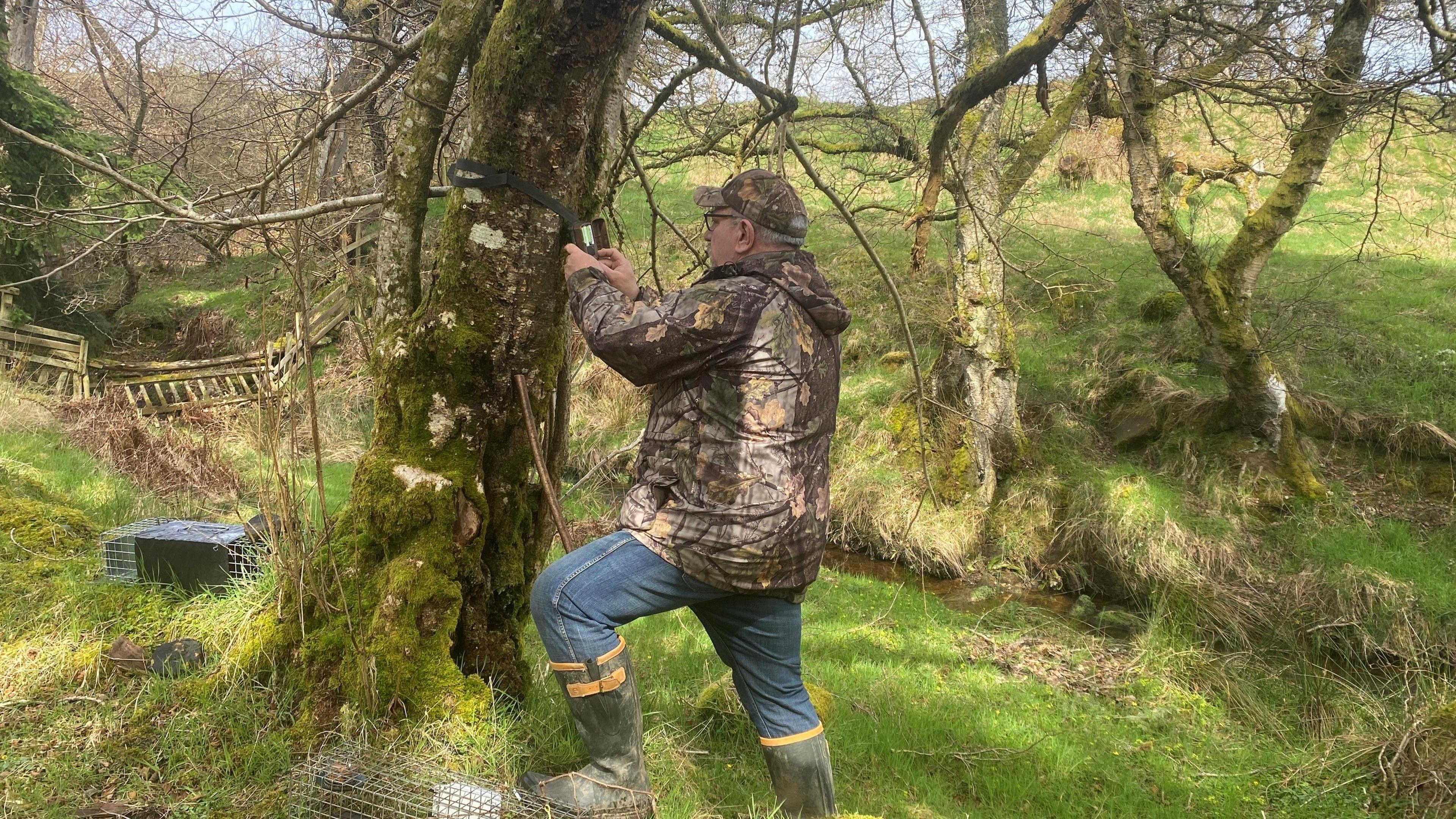 Picture shows Ian Glendinning putting up some video equipment in a woodland setting. There is a trap on the ground.