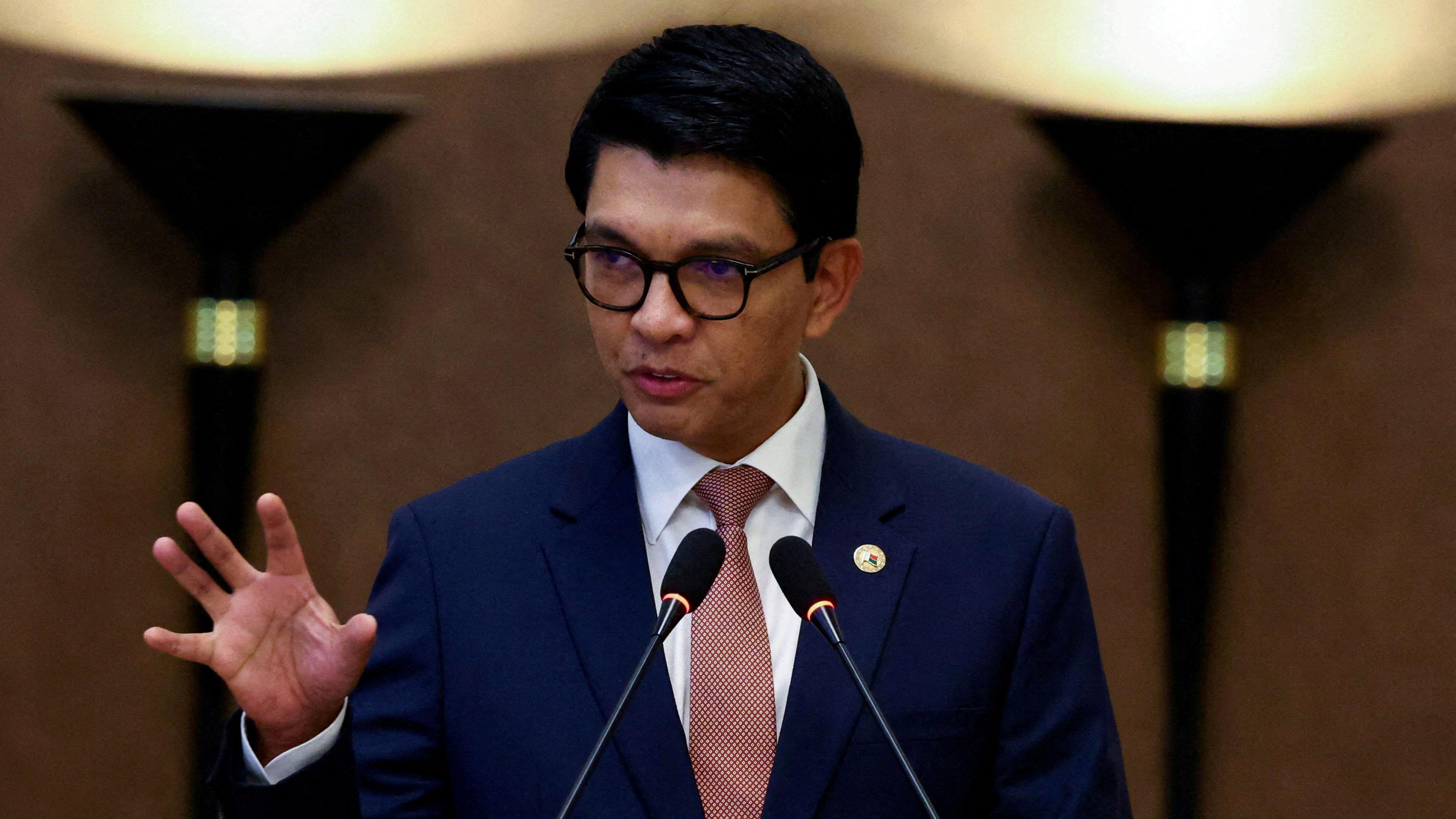 A man wearing black-rimmed glasses and in a blue suit with a red tie speaking into two microphones, holding up his hand as he speaks