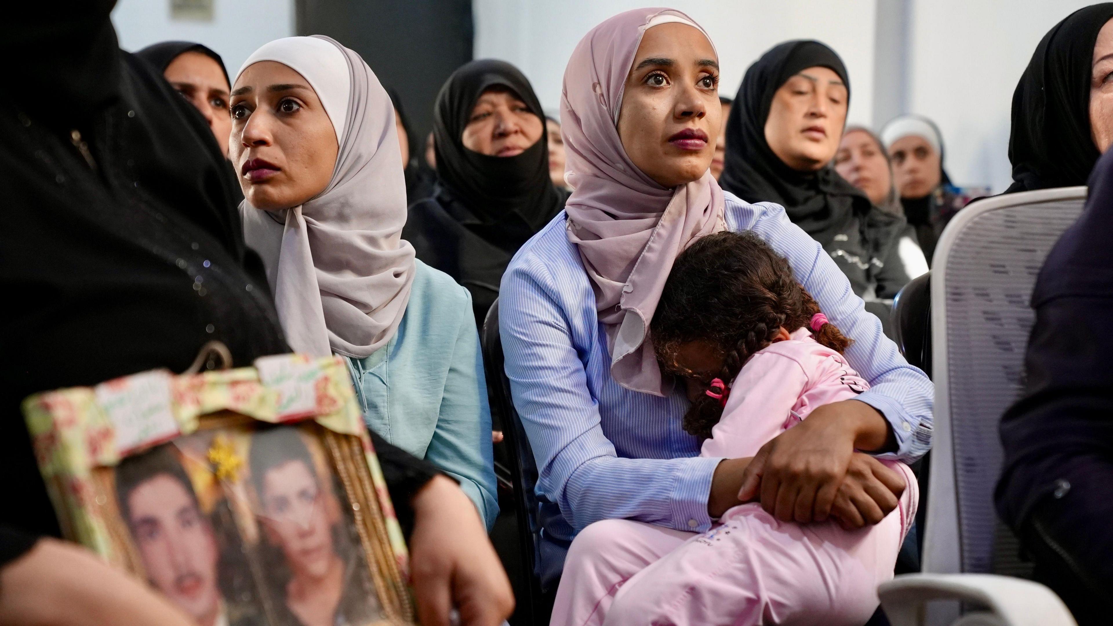 A young mother sits in a hall, her young daughter on her lap. Around her sit other women, many older, wearing black robes.