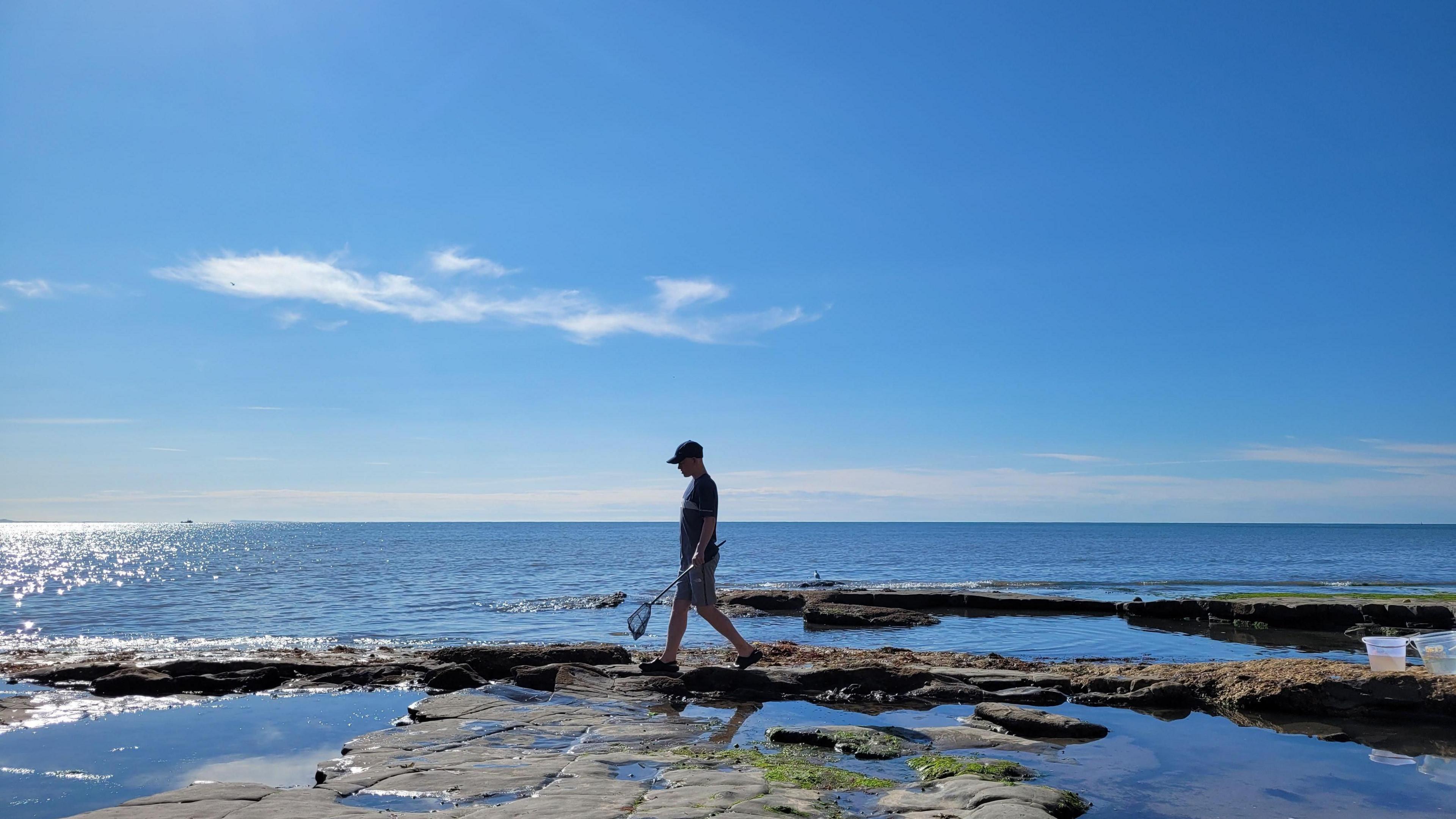 A man wearing shorts, tshirts and a cap walks across rocks carrying a small net. The rocks are on the edge of the sea beyond that is bright blue in the sun. He is wearing black shoes and there are several rock pools. The sky is blue with one fluffy white cloud. 