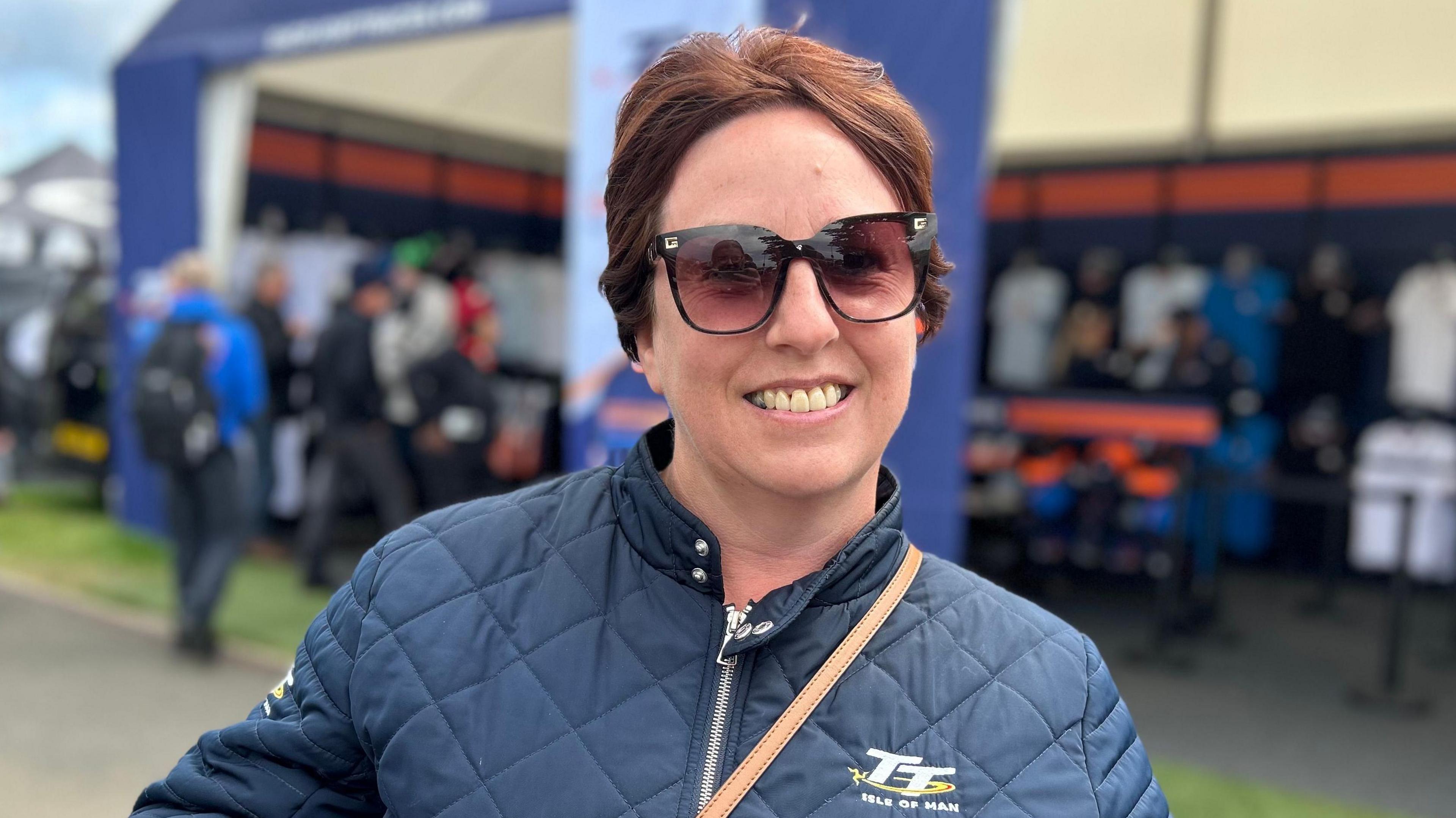 A woman in a TT jacket wearing sun glasses and with reddish hair standing in front of the TT merch stand