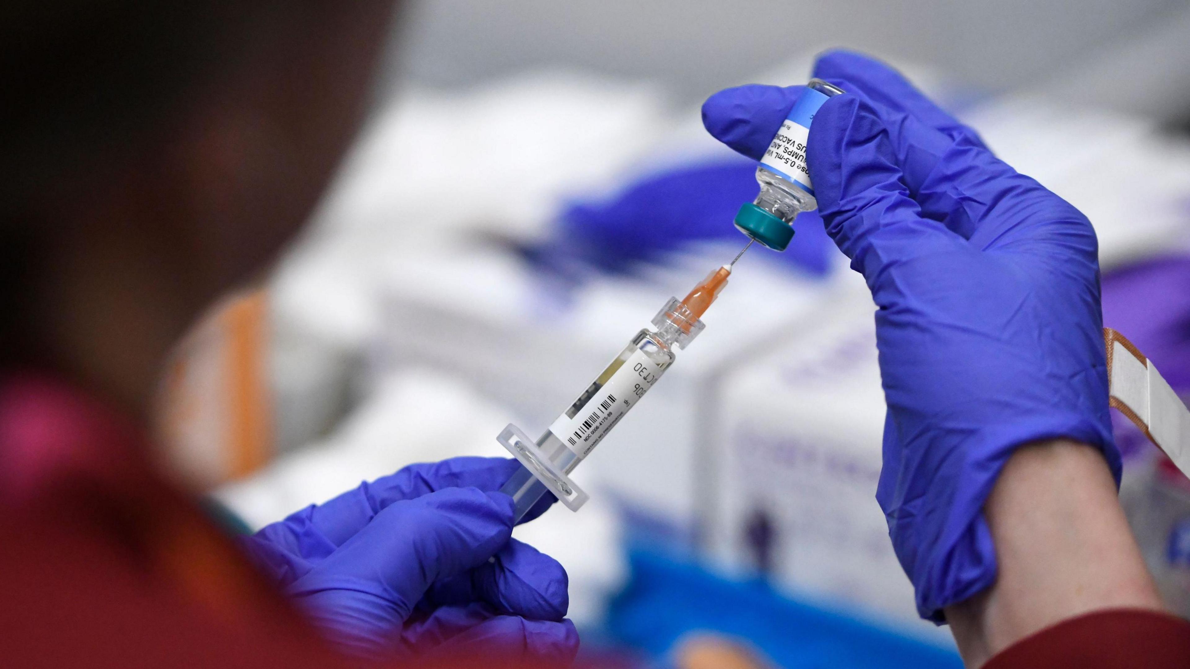 A medical staff member wearing blue gloves uses a hypodermic needle to take medicine out of a small glass vial