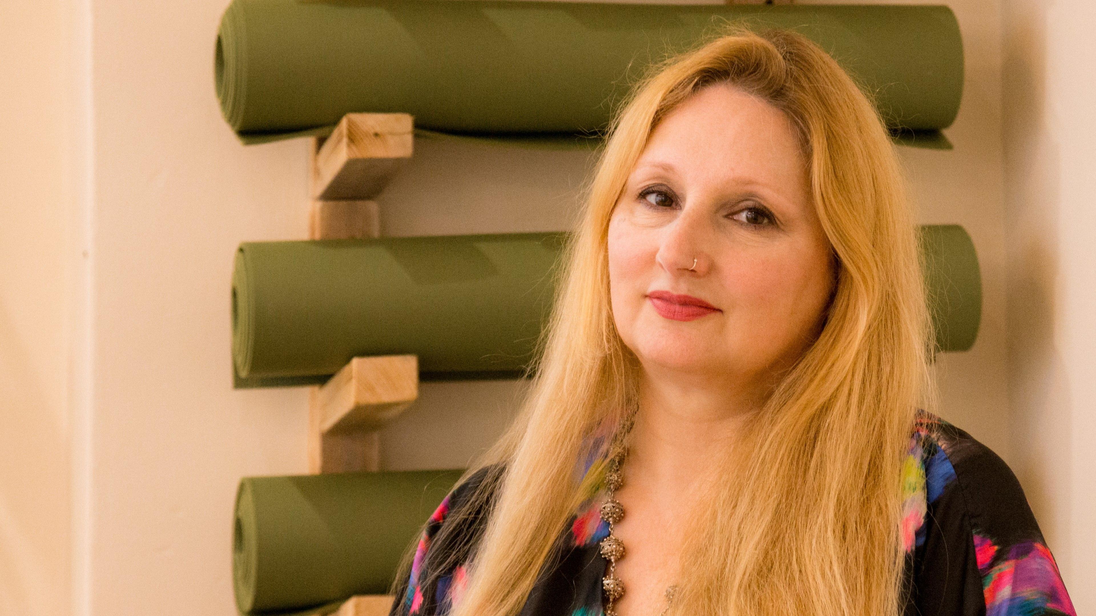 Florence Achery has long blond hair and is standing in front of a rack of yoga mats.