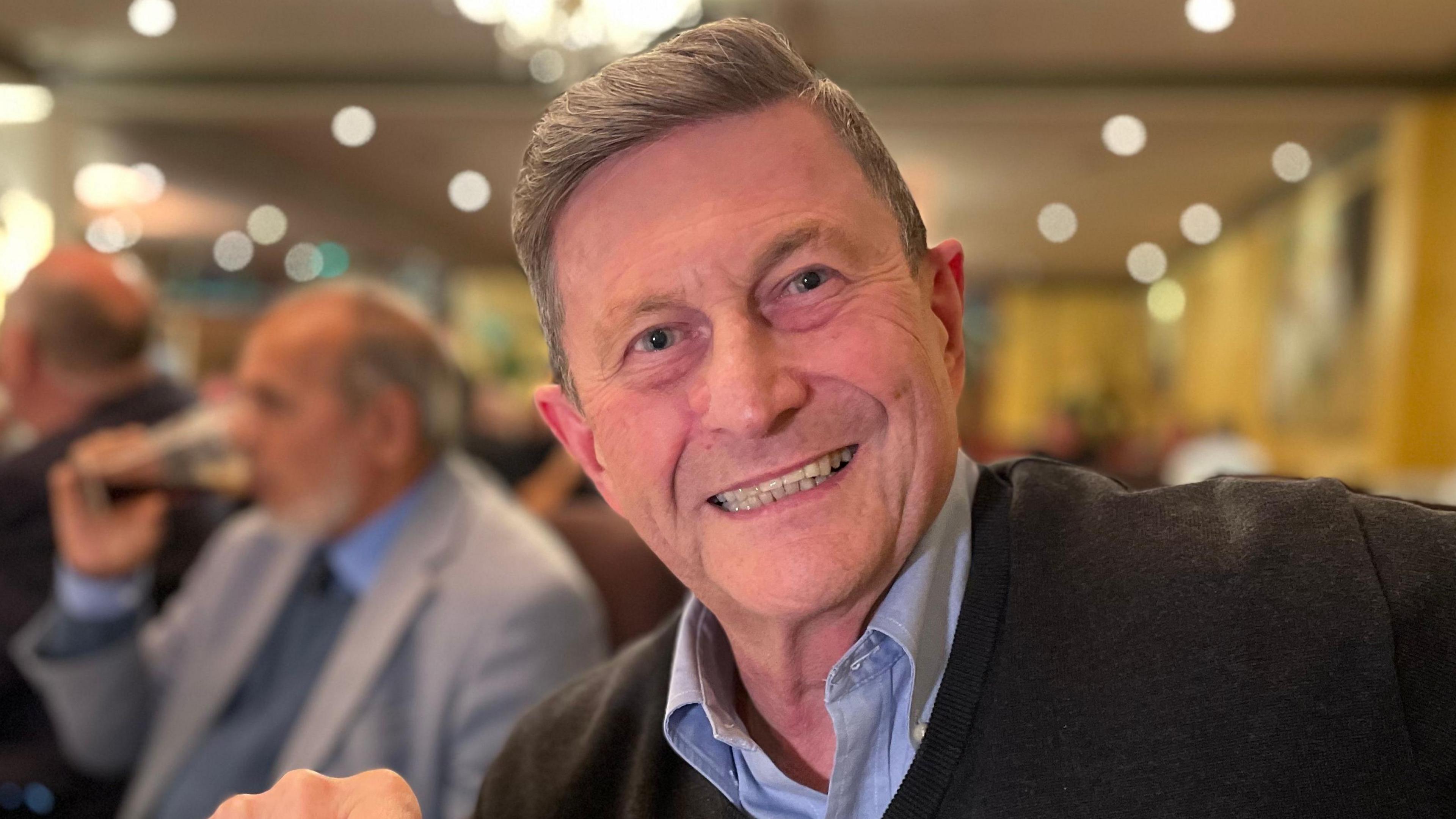 Steve Allen smiling and looking direct to camera. He is wearing a dark coloured v-neck sweater over a light blue shirt. He has mousy coloured hair and blue eyes. Other diners in a restaurant can be seen out of focus behind him.