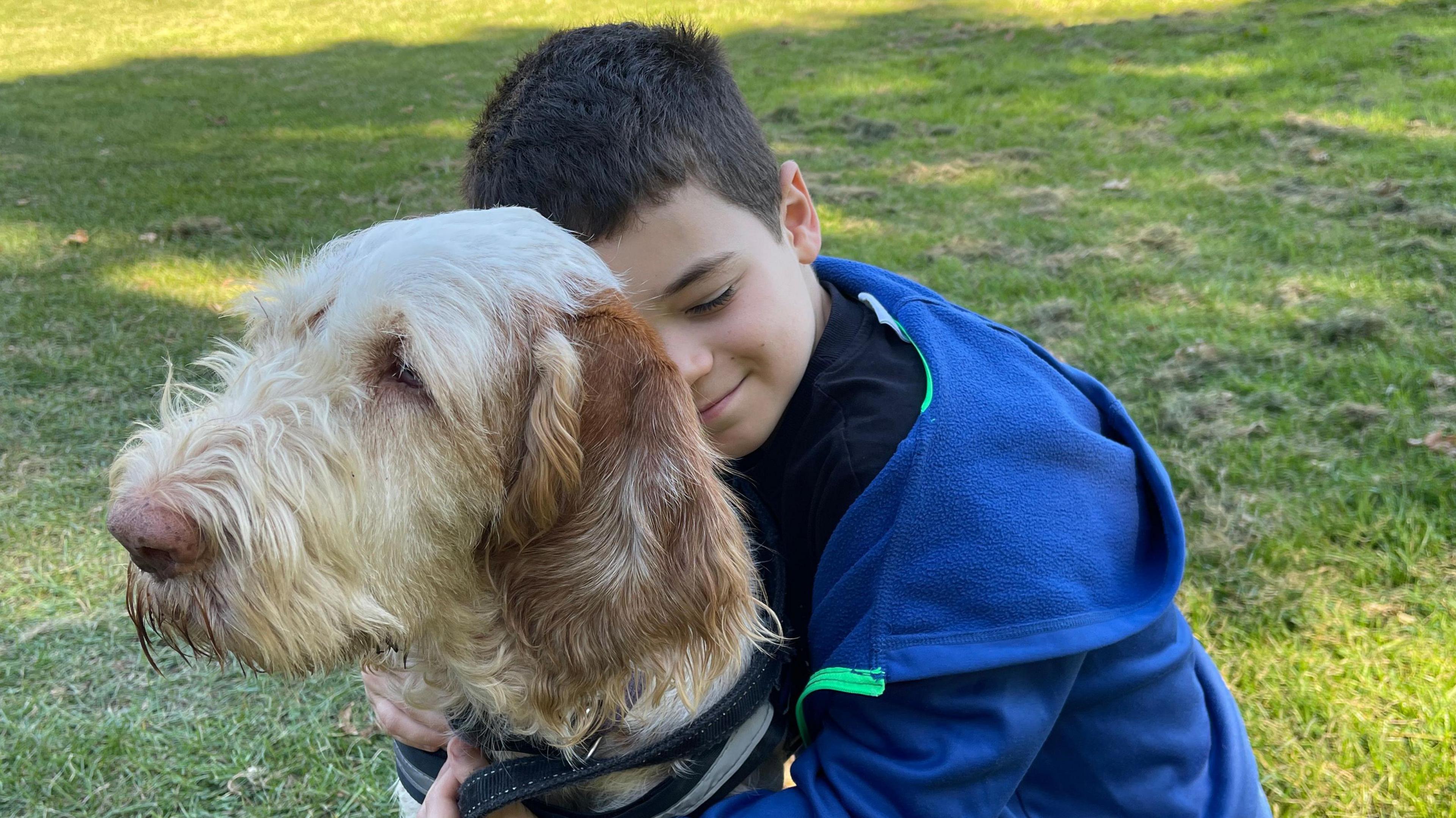 Ten-year-old Isaac Thompson, who as a a dark short back and side hair style and is wearing a blue hoodie with bright green piping down the zip line, hugs Hero from behind