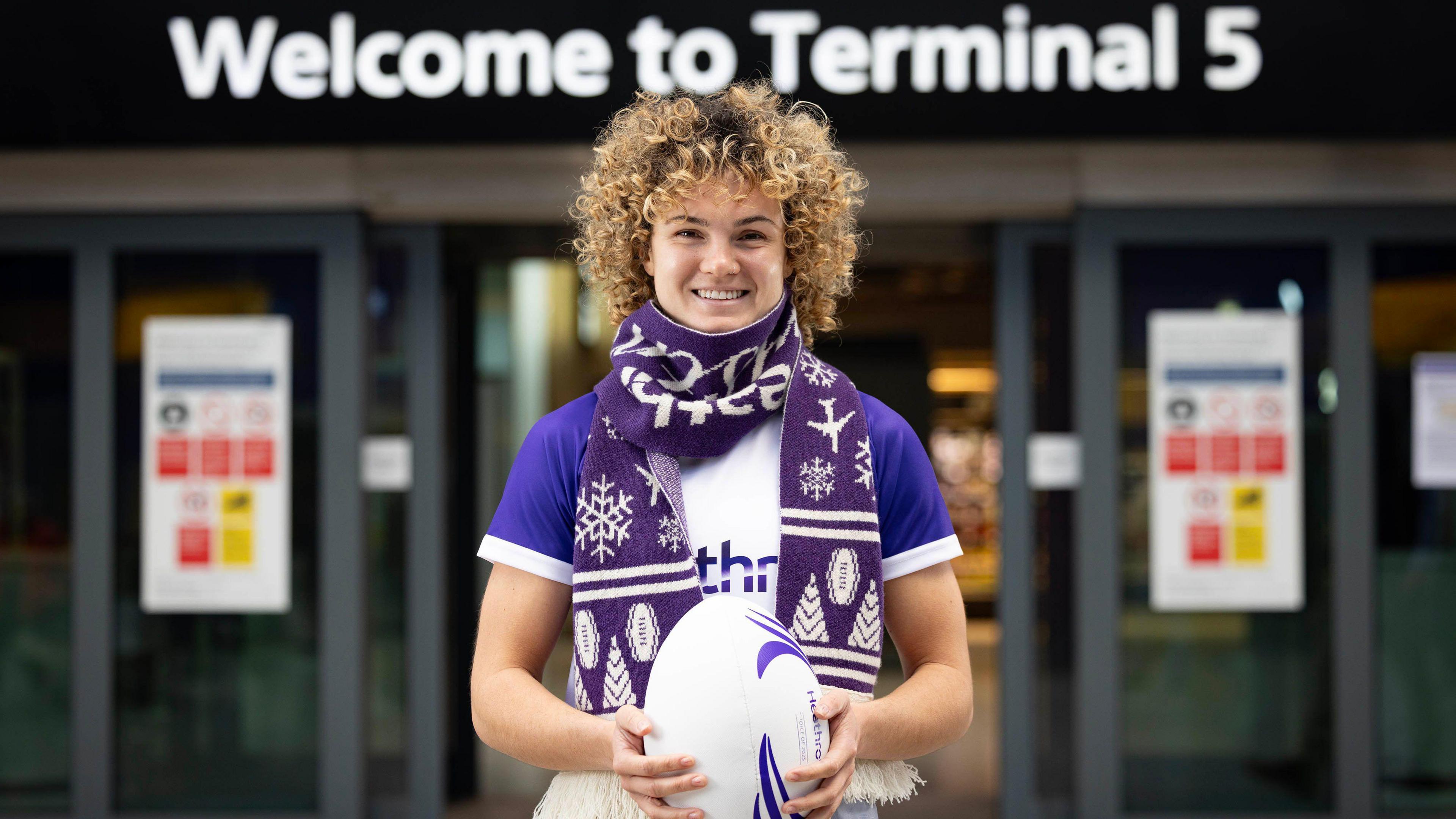 Ellie Kildunne standing in front of Terminal 5 at Heathrow airport holding a rugby ball, smiling straight into the camera, wearing a thick blue Christmas skarf