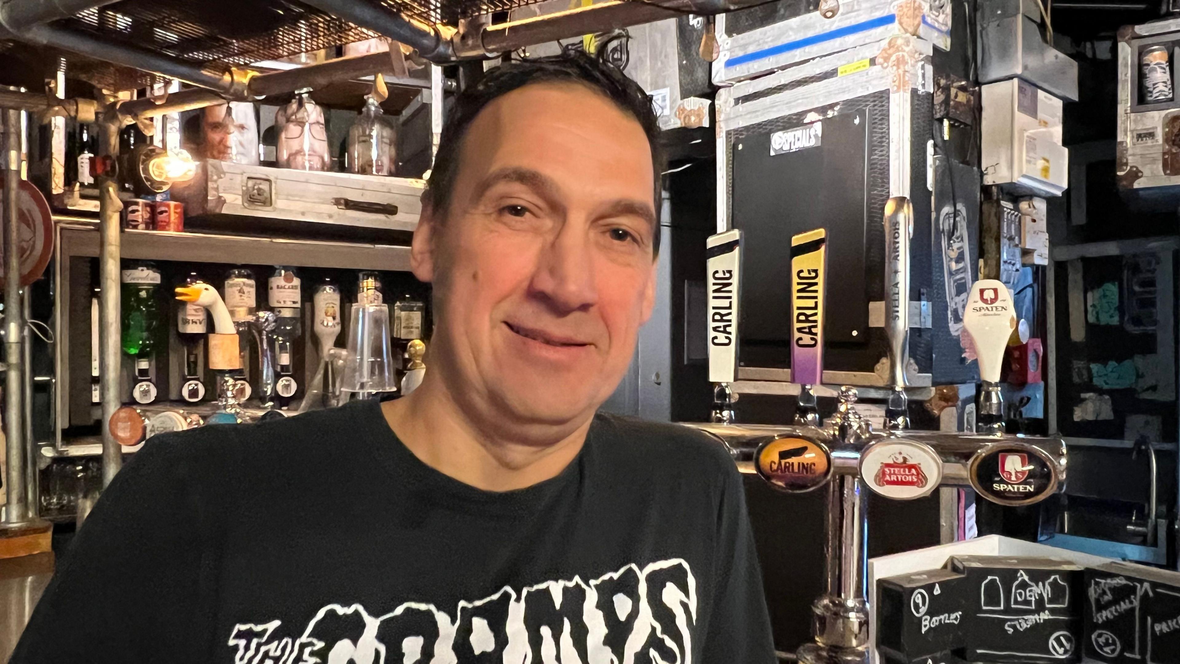 Chris is wearing a black t-shirt with The Cramps emblazoned on it. He is standing at a stocked bar made of metal black and grey boxes.