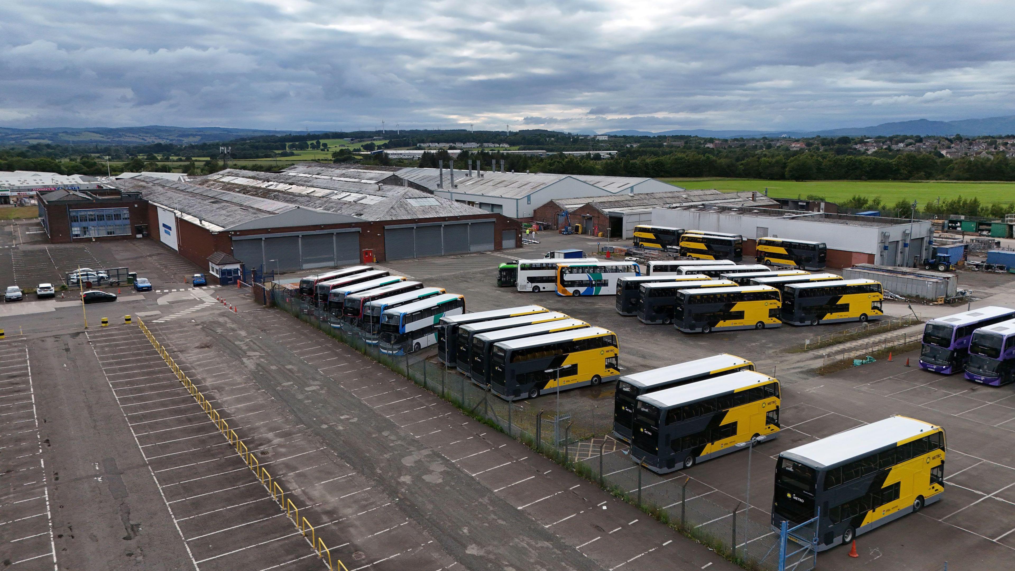A general overview of the Alexander Dennis site in Camelon. A number of yellow and black buses are parked in a car park on the right side while an empty car park is on the left side.