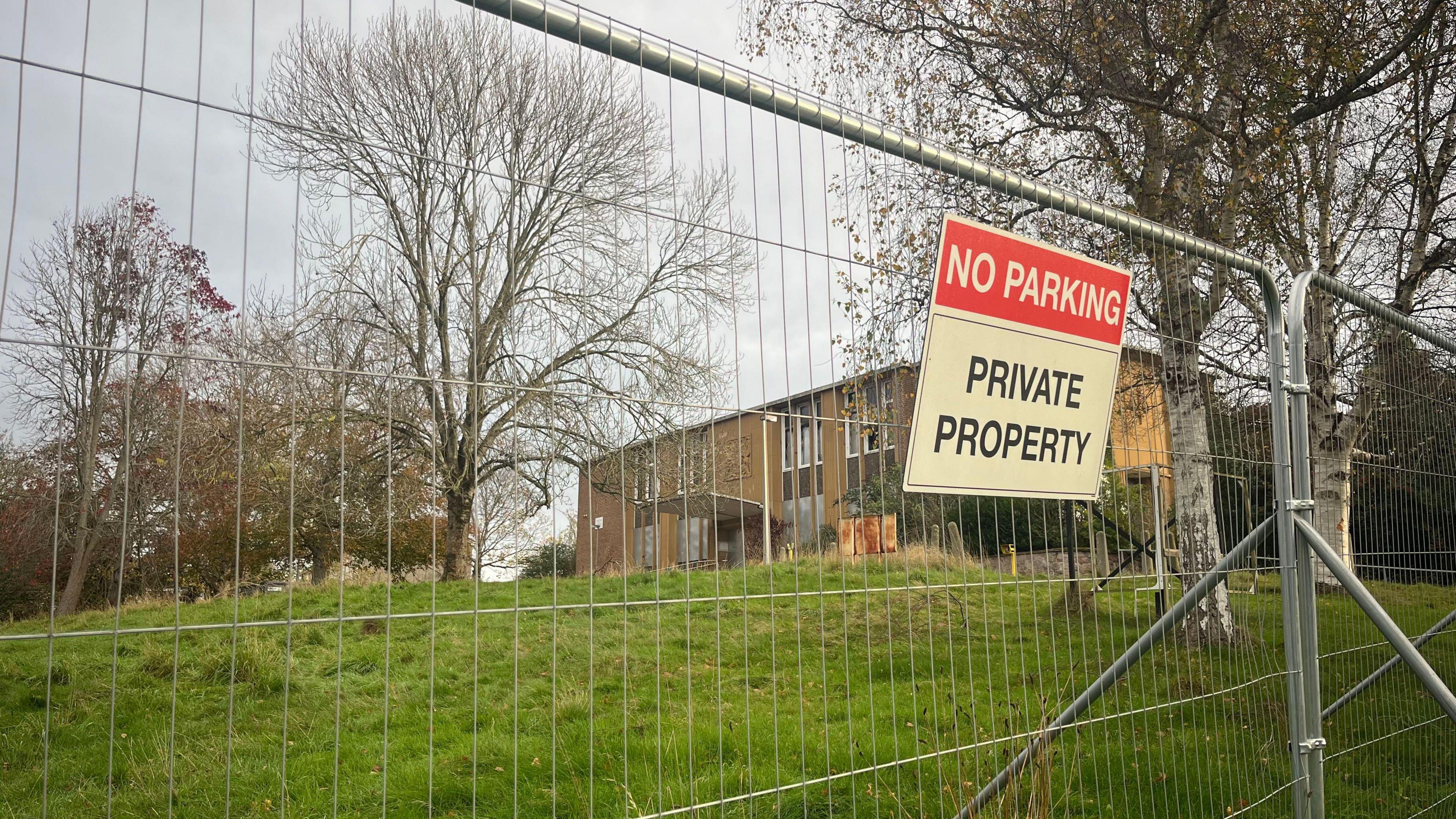 A metal barrier in front of the old police station building. A no parking private property is displayed on the fence. A grass bank leads up to the property. 