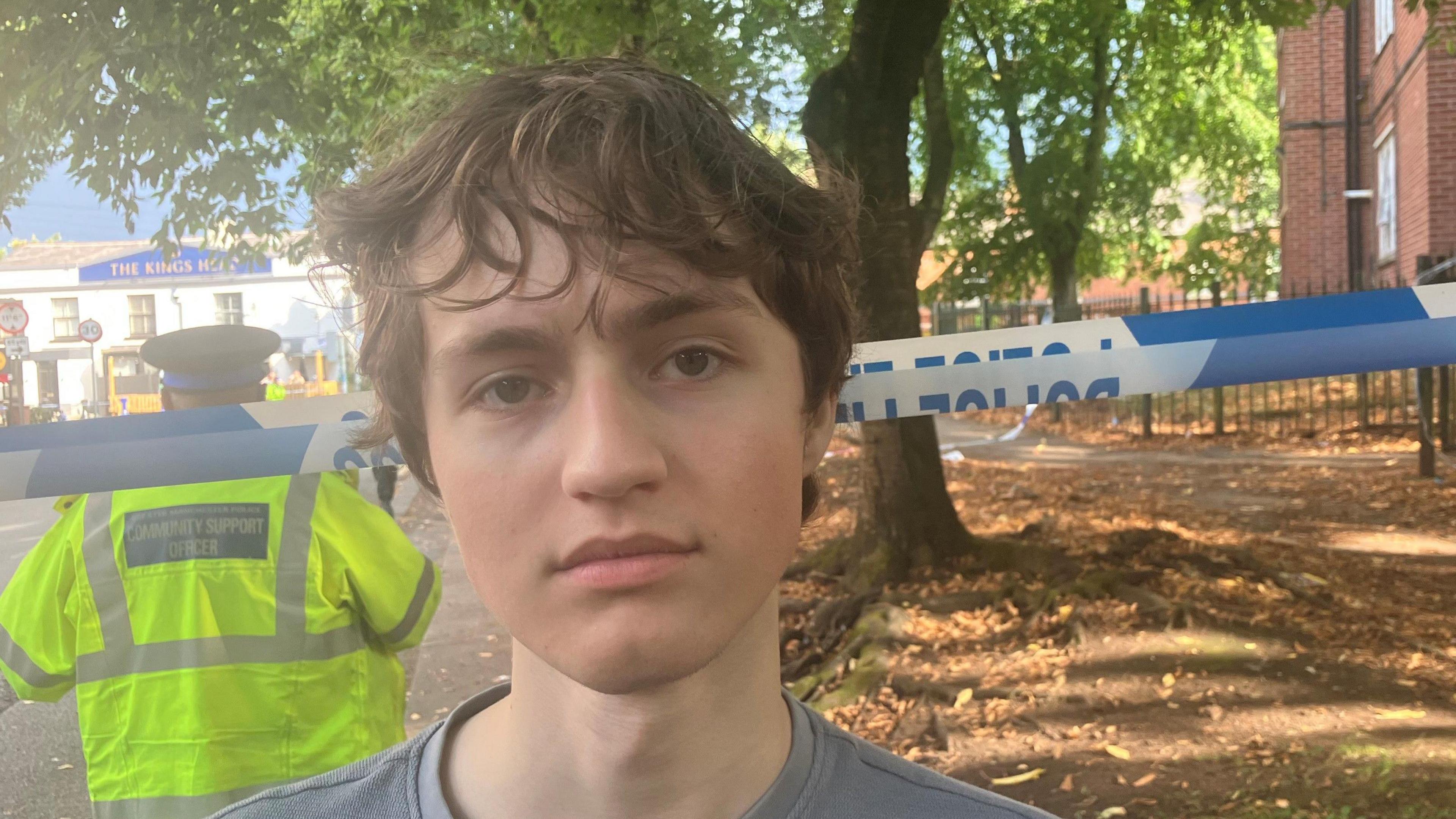 A young man with messy light brown hair stands with a neutral expression in front of police tape which is across an area in front of a tree. A police officer is stood facing away behind him.