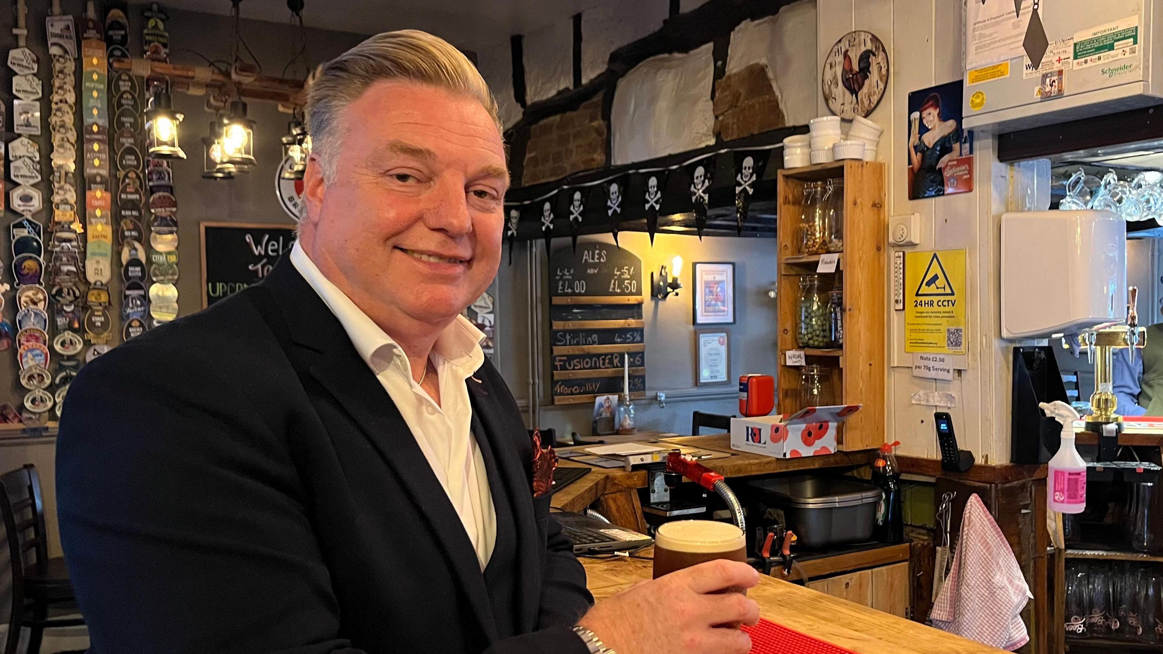 A smiling Marvin Shalders looks directly at the camera, wearing a white shirt and dark suit jacket, holding a pint of beer at the pub bar.