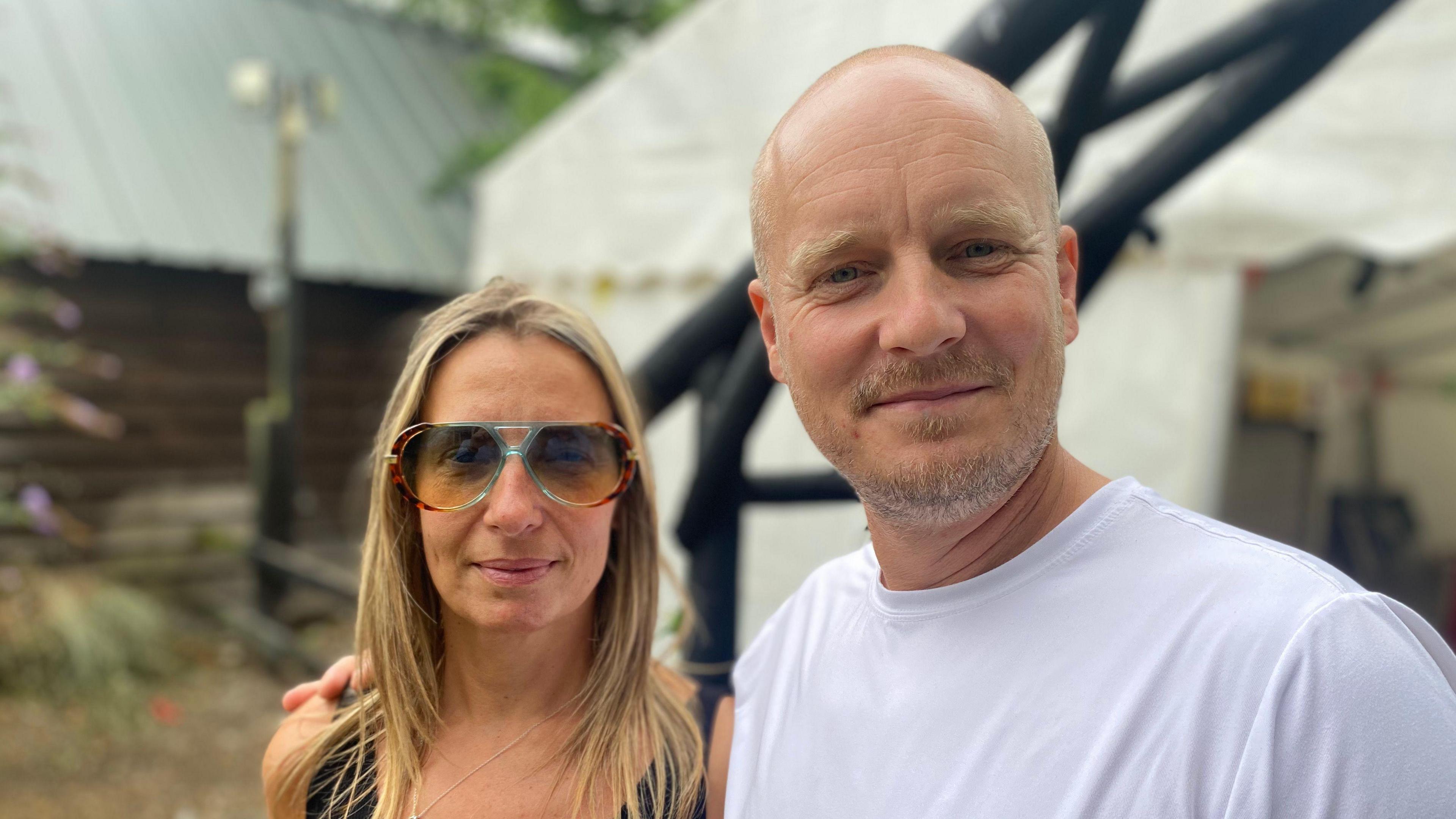 Mr and Mrs Dixon are smiling. He has his right arm around her shoulders. Mr Dixon has a shaved head and some facial hair and is wearing a white top. Mrs Dixon is wearing sunglasses and has long blonde hair. They are standing in front of a white marquee.