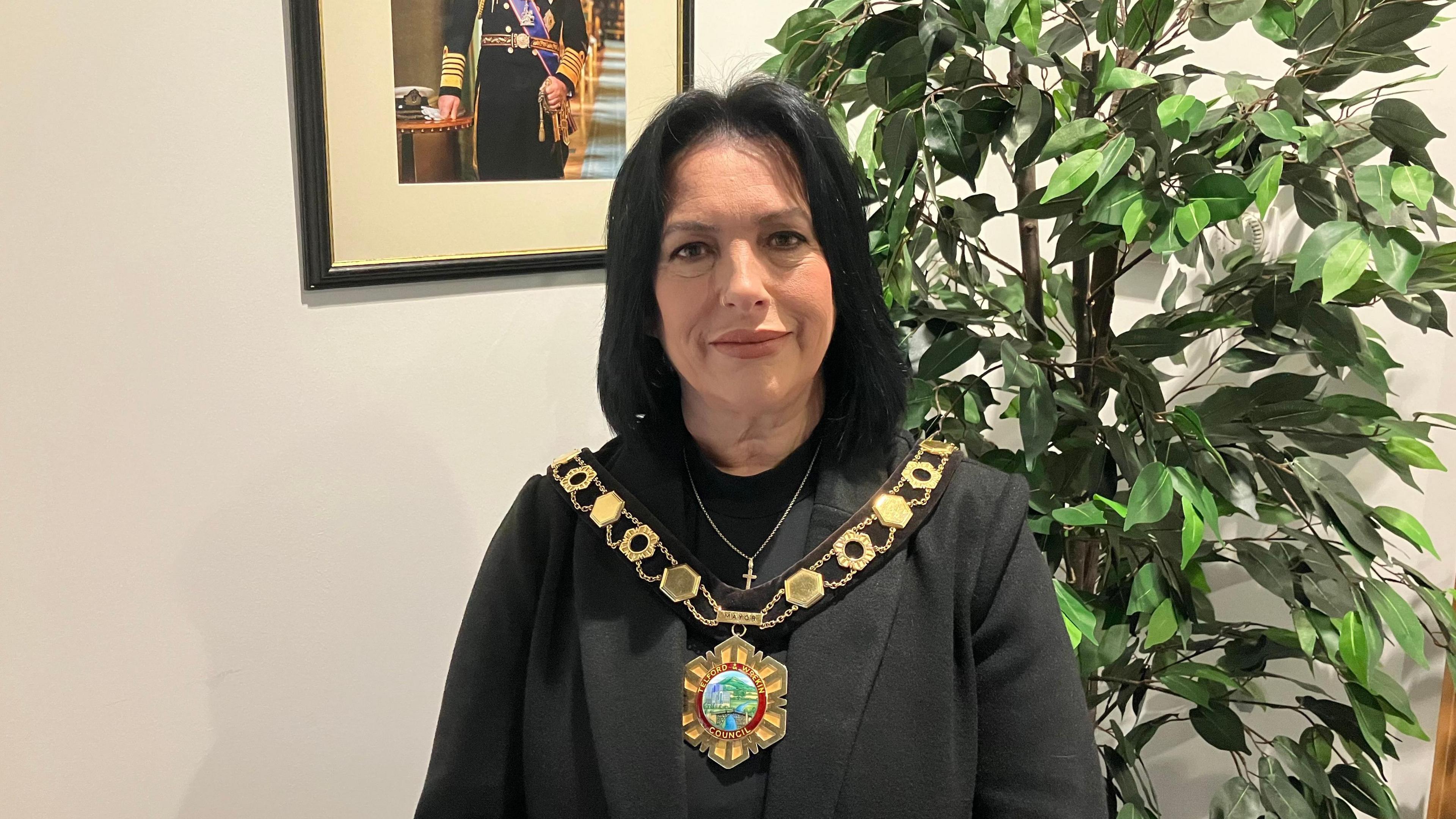 A woman with long dark hair and a dark top with a gold chain of office around her neck and a green plant behind her