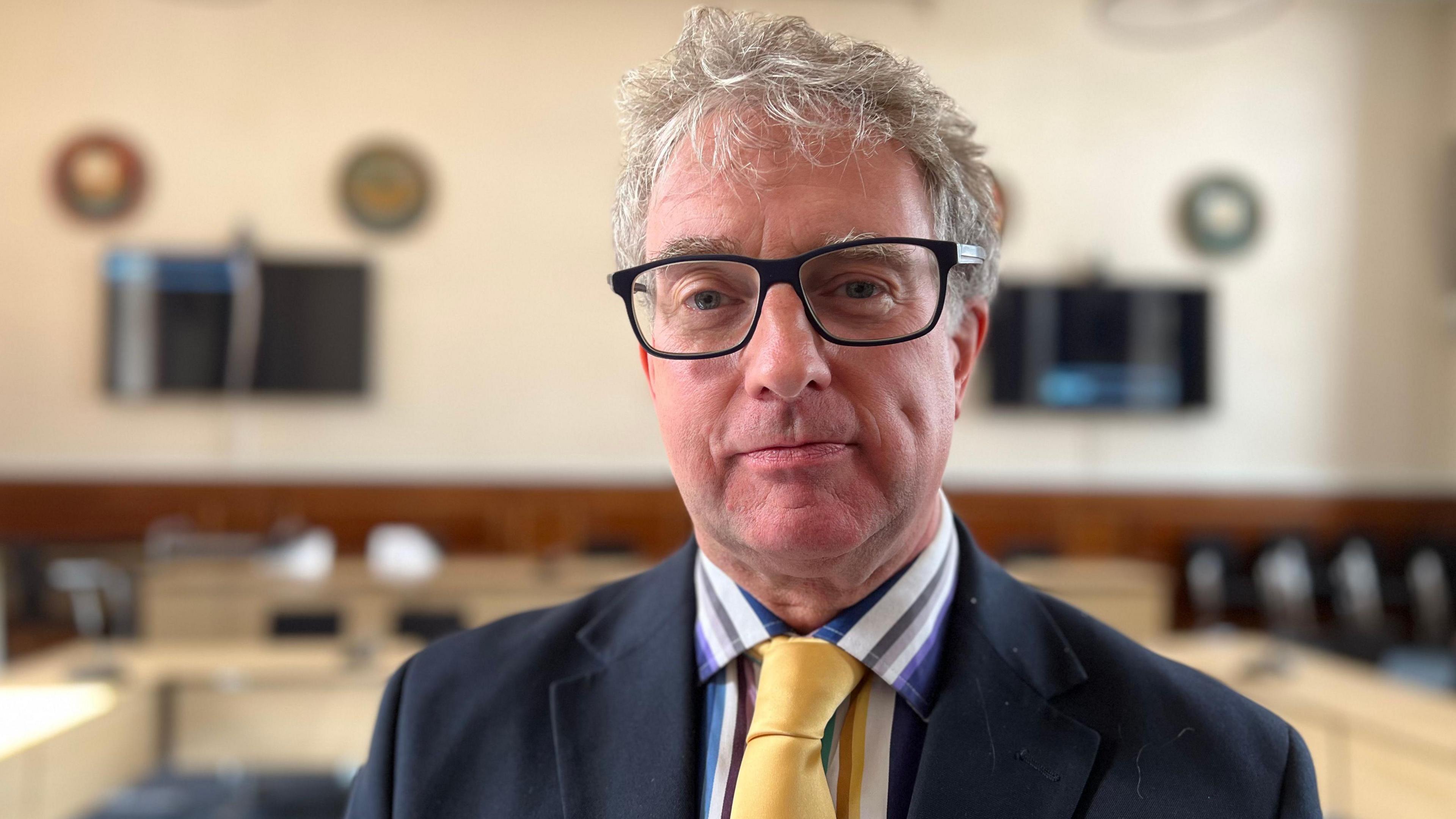 Head and shoulders picture of Jonathan Bacon in the council chamber, wearing a dark blue jacket, striped shirt and yellow tie 