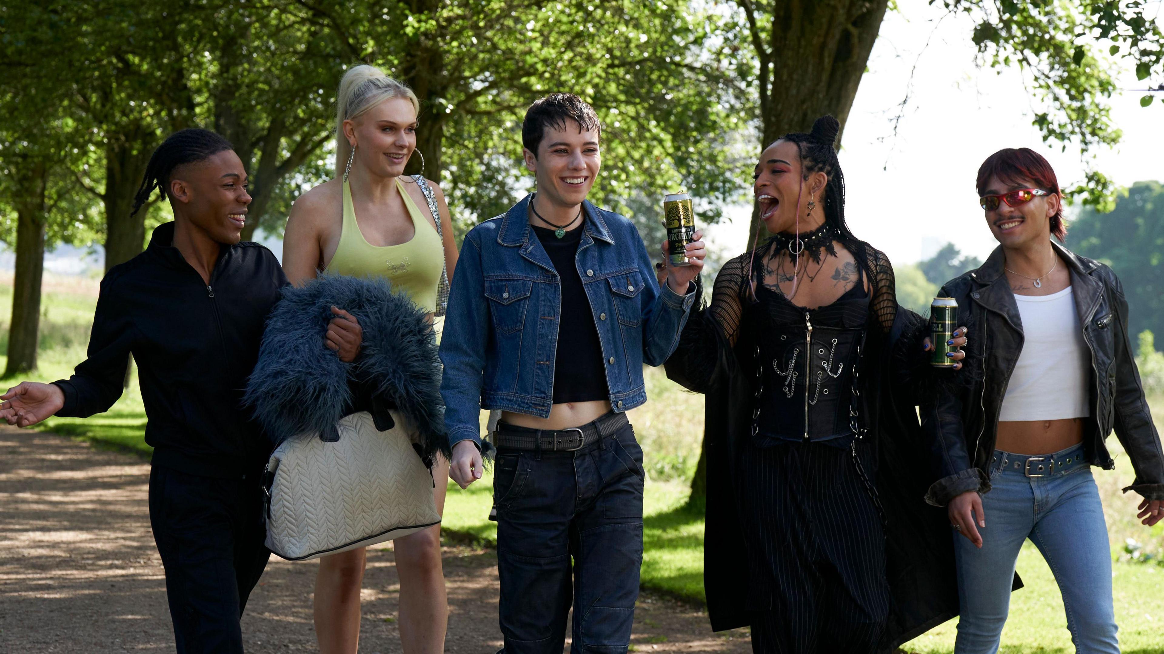 Characters of What It Feels Like For A Girl are walking along a path in park. Ellis Howard playing character Byron is in the centre of the group wearing a denim blue jacket over a black top and wearing blue jeans. 