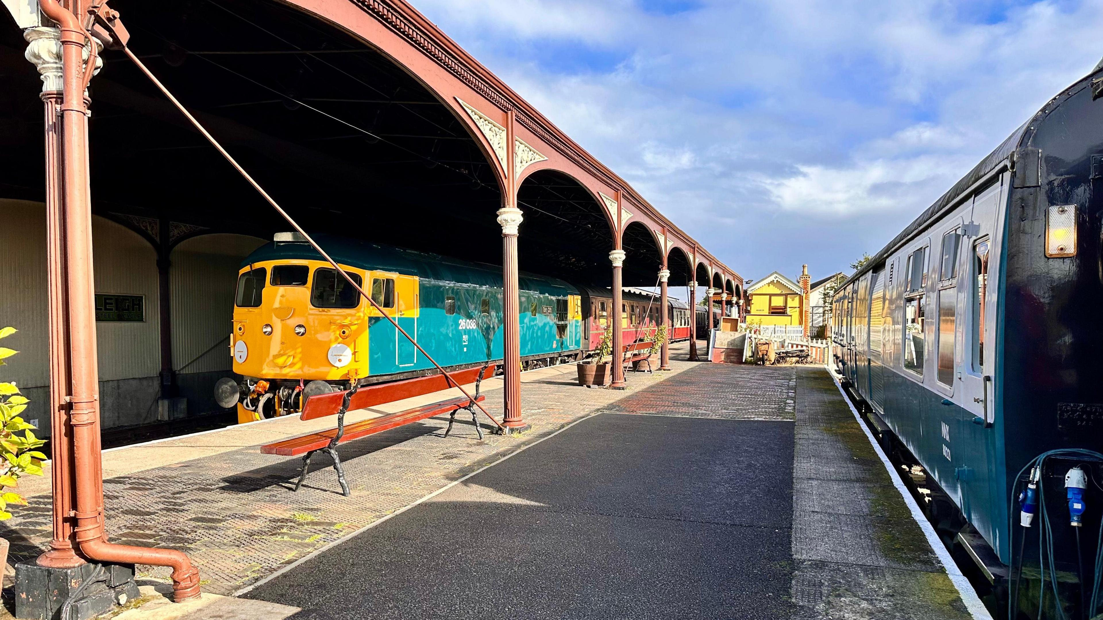 Trains in a station with the one on the left looking brilliant bright yellow and green in the sunshine.