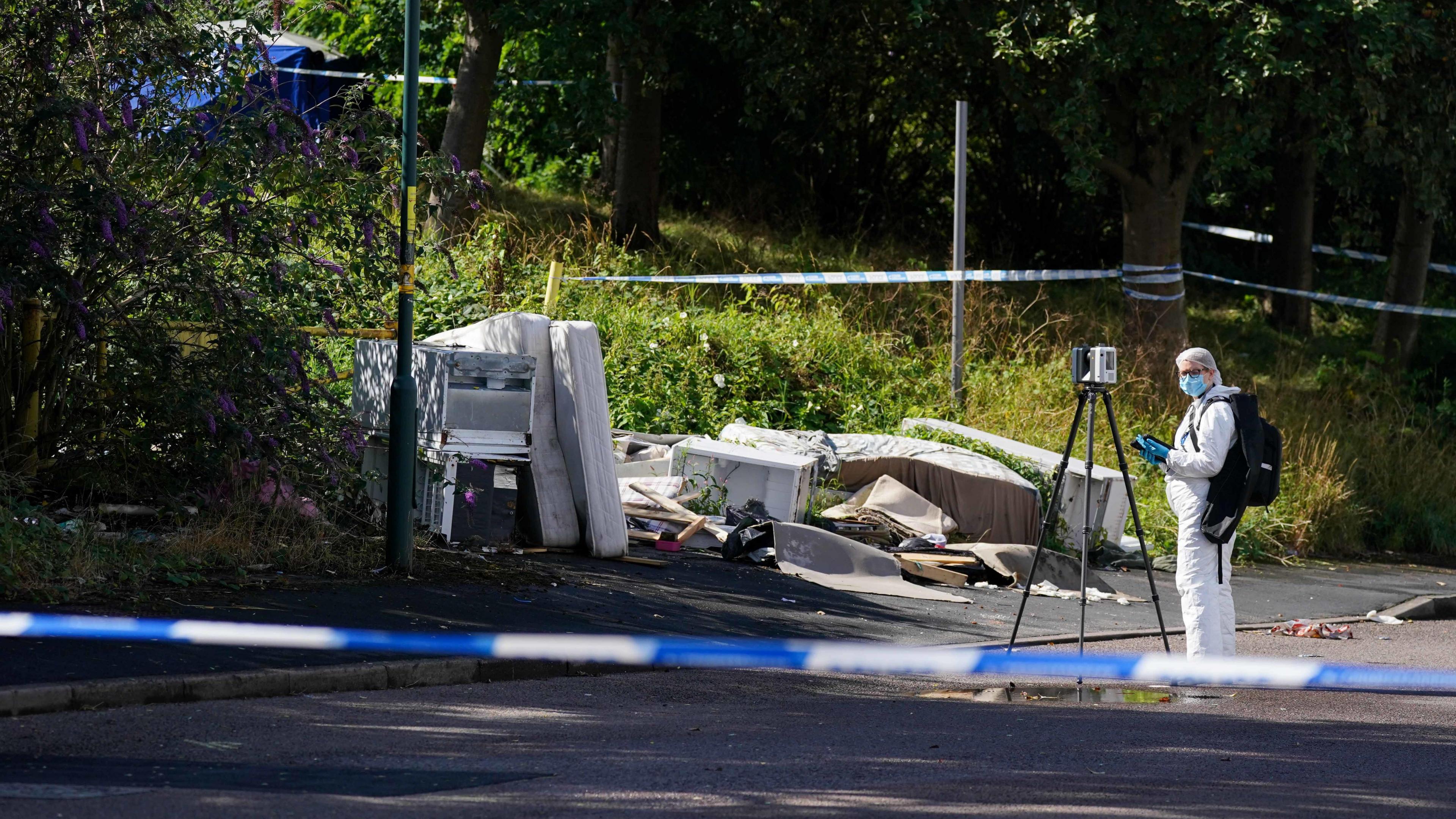 A man in a white forensic suit stood in front of a tripod and camera. He is at the scene of a woodland area. There are various bits of old furniture scattered. The area is cordoned off with police tape. 