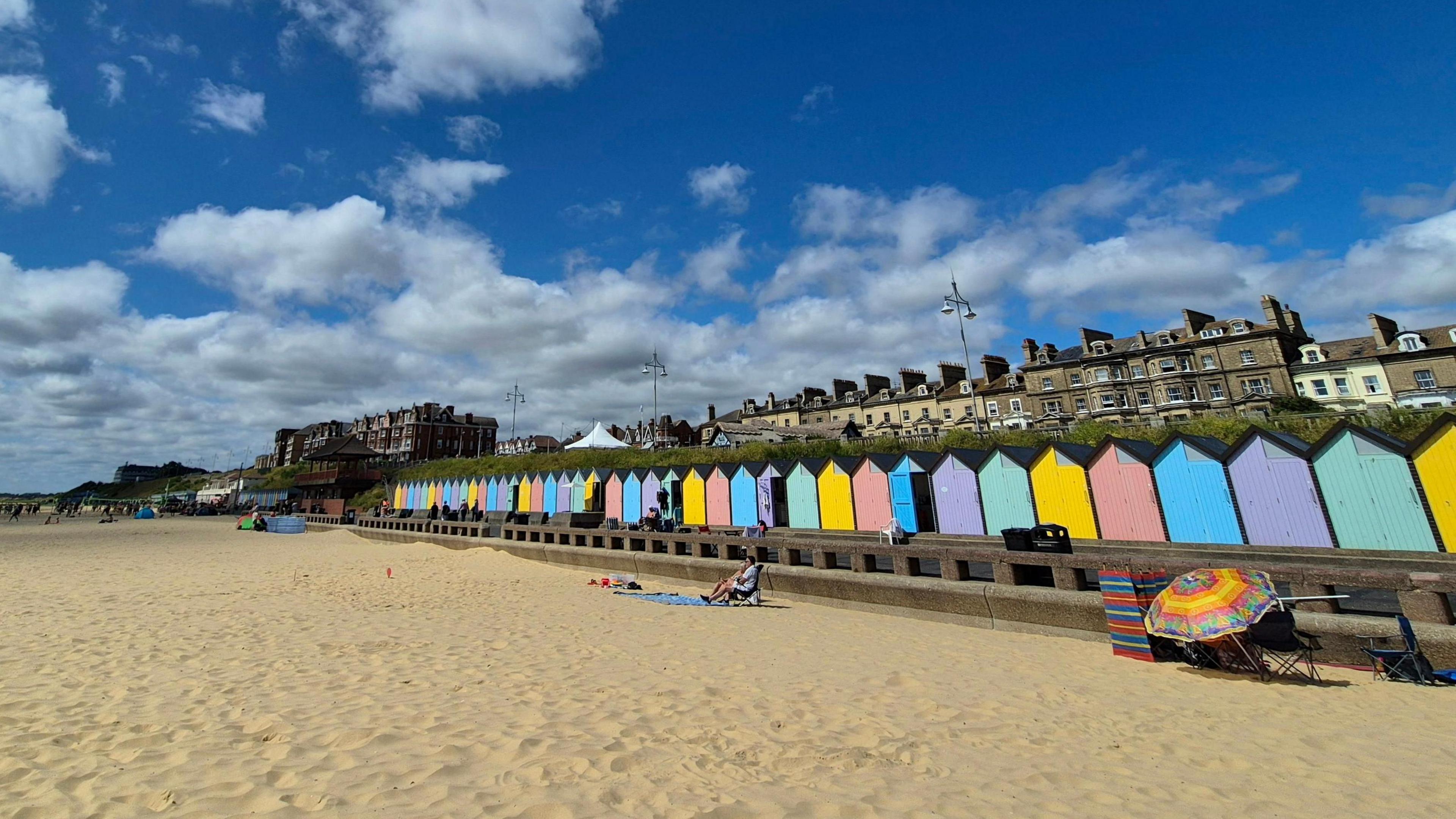 A view standing on the beach looking towards the beach huts and houses on the sea front. The beach huts are different colours, and two people are sitting in front of them on the sand in chairs. 