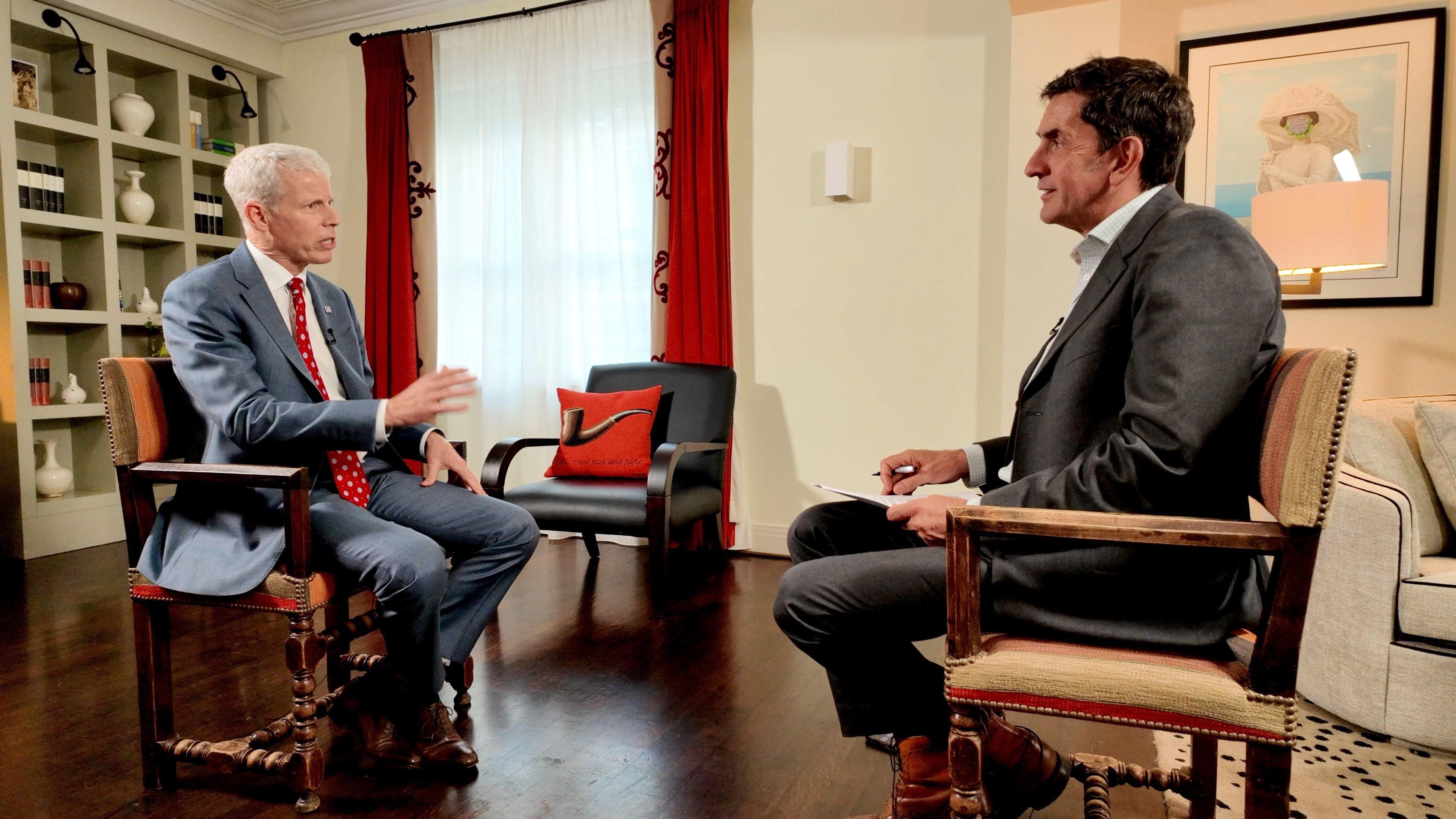 Chris Wright is on the left and Justin Rowlatt is on the right. They sit facing each other in what looks like a study on wooden armchairs. Wright wears a grey suit with white tie and red shirt and Rowlatt wears a grey suit.