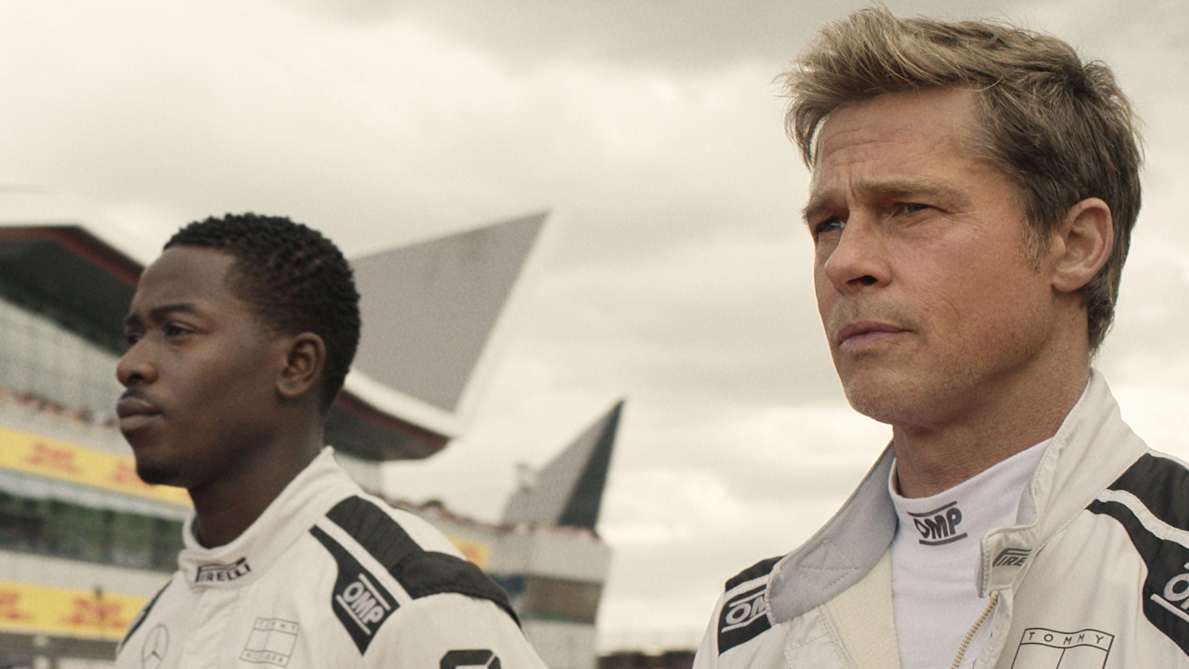 Damson Idris as Joshua Pearce and Brad Pitt as Sonny Hayes' headshots. They are seen from the side, walking and looking to the left of the shot with serious expressions. They wear white F1 jackets with OMP printed on the shoulder. To their left is the stadium.