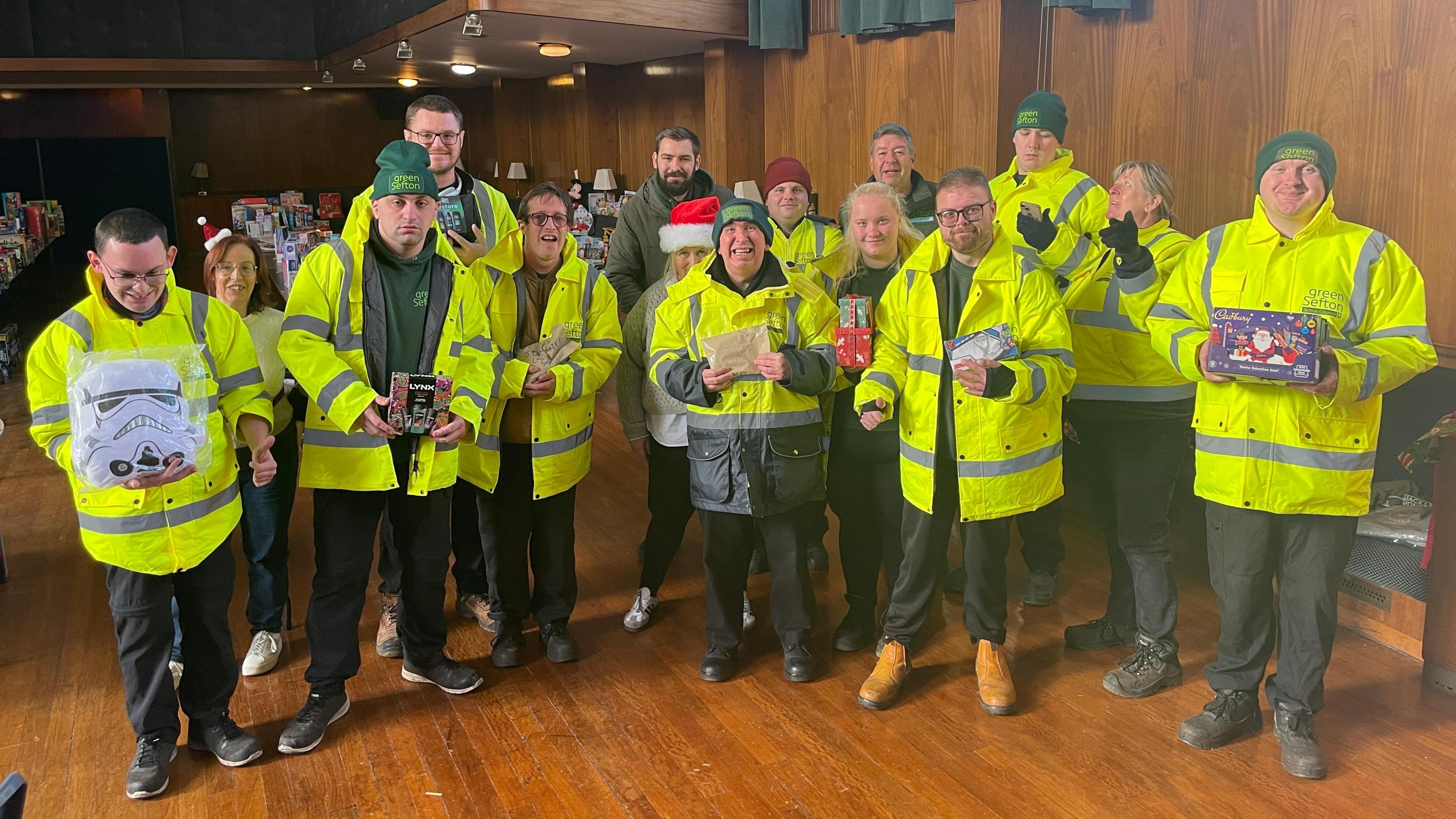 A large group of people wearing high viz yellow coats holding a selection of toys to camera and smiling ins ide Bootle Town Hall