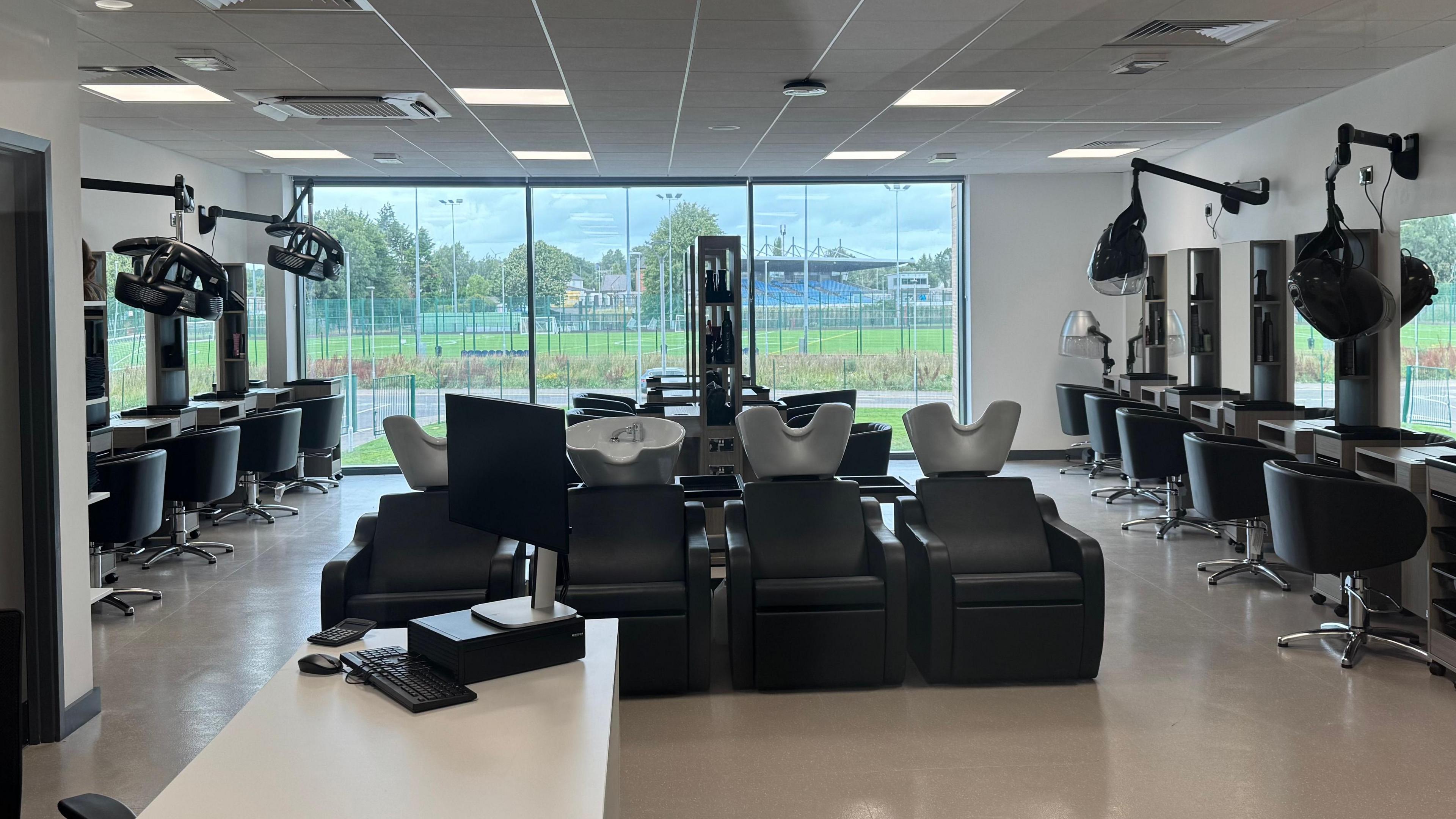 A large white room with floor to ceiling windows at the far end. Around the walls of the room are hair-dressing stations with chairs, mirrors and shelves for equipment. In the centre of the room are a number of hair washing stations with sinks and black leather-like recliner chairs.