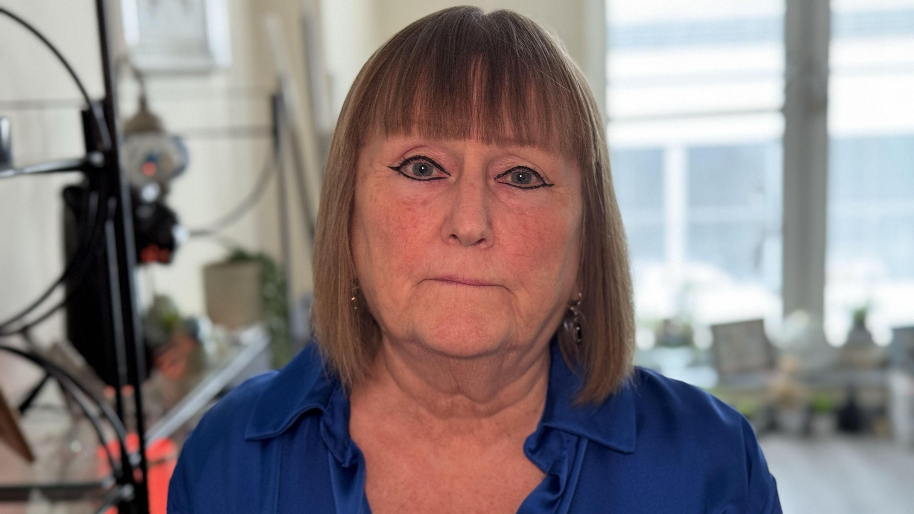 A woman with brown hair and a blue shirt, stands in the living room of an apartment.