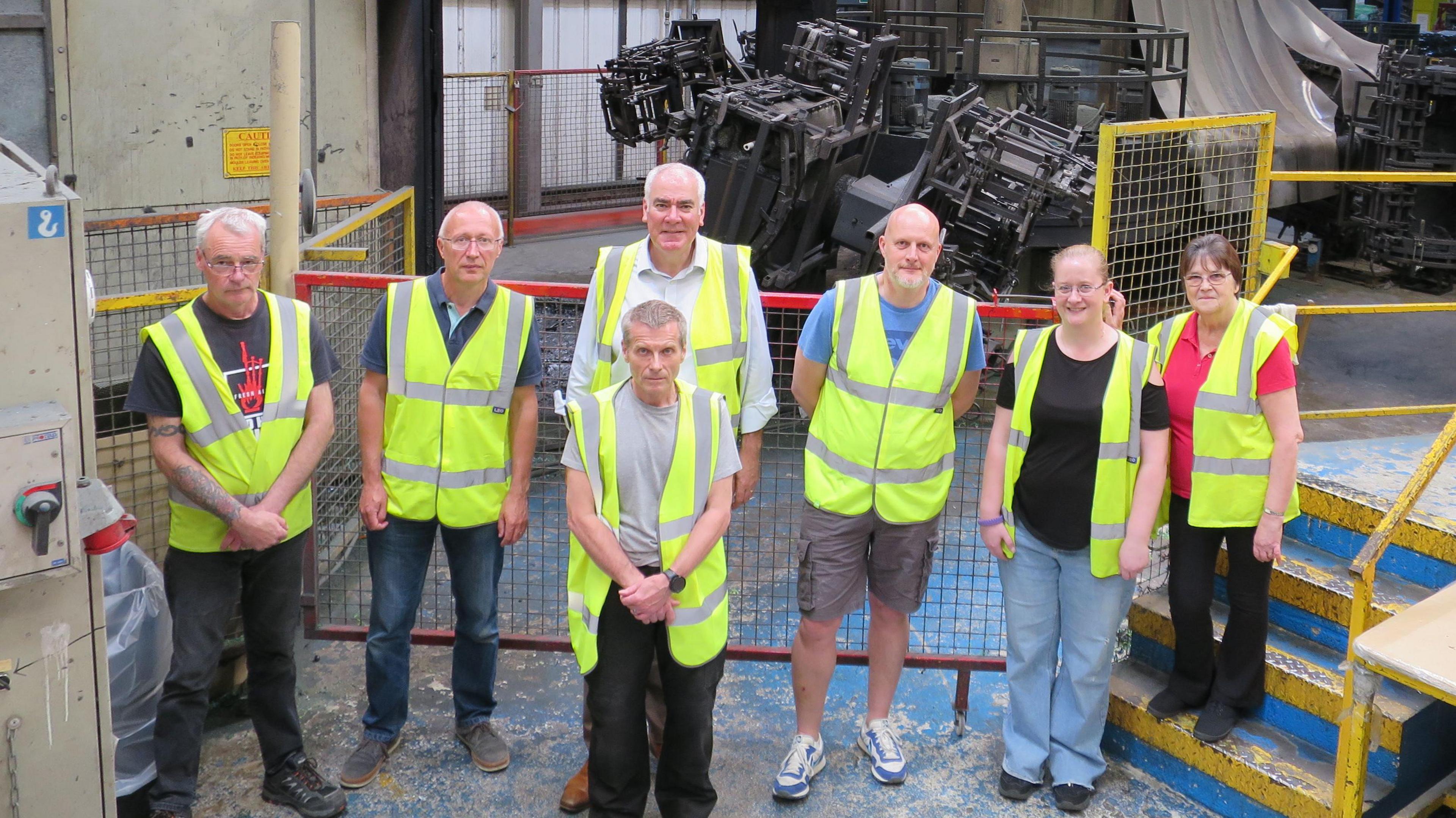 A group of staff in high-vis in a factory all looking up at the camera. Complicated machinery and pipework behind them.