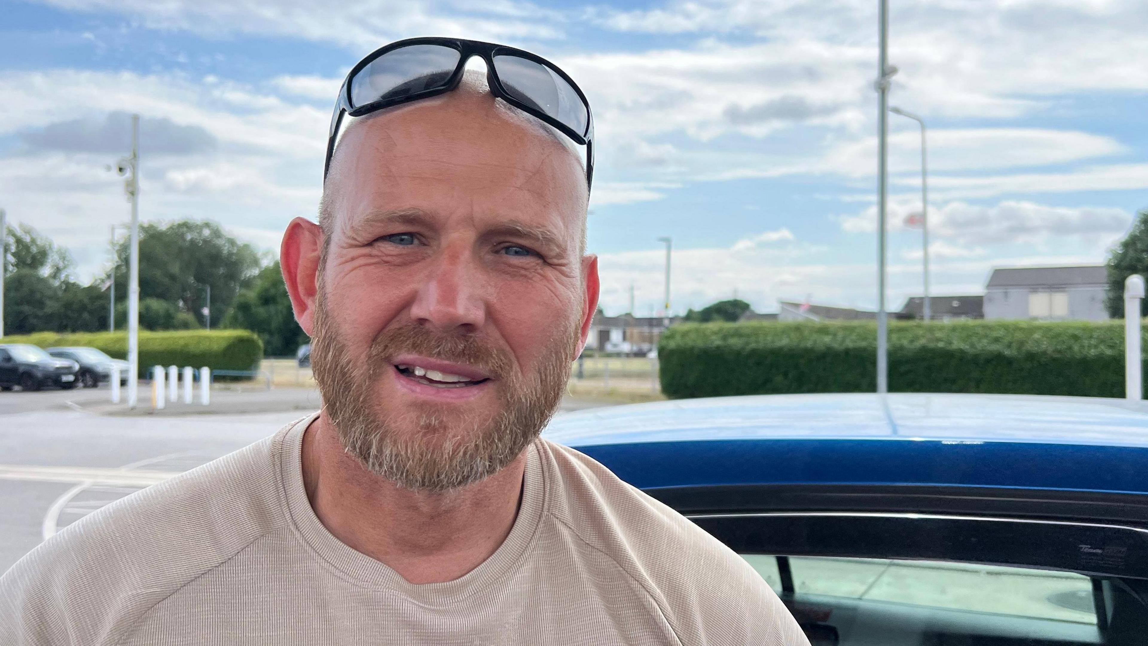 Wayne Parsons is a muscled and bald middle aged man with blue eyes and sunglasses on his head. He's wearing an immaculately laundered beige T-shirt and is standing beside his ultramarine car at a petrol station.