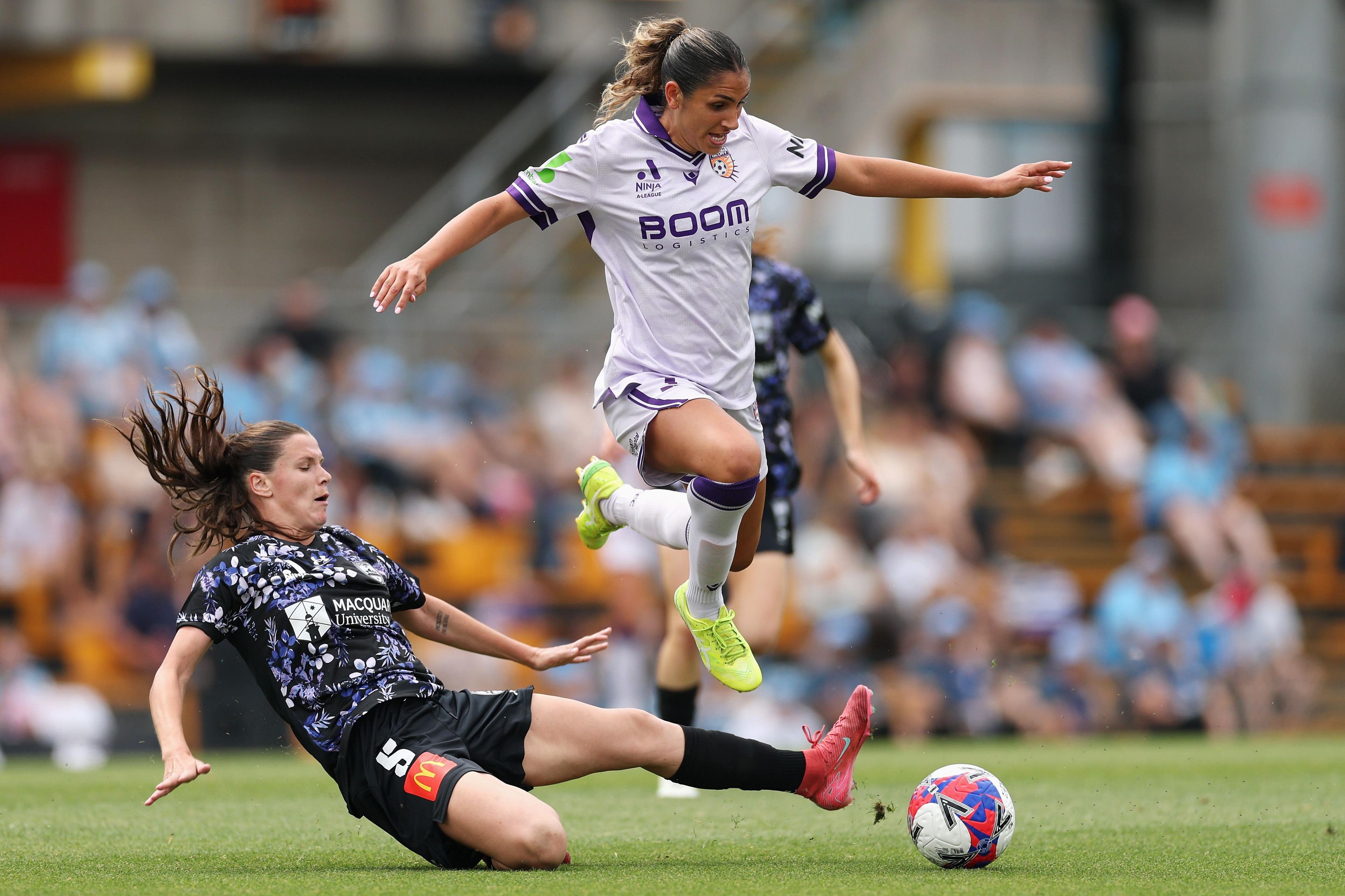 Soccer player in a white and purple kit leaping over an opponent in a black patterned kit attempting a sliding tackle, with a colorful ball on the grass and blurred spectators in the background.