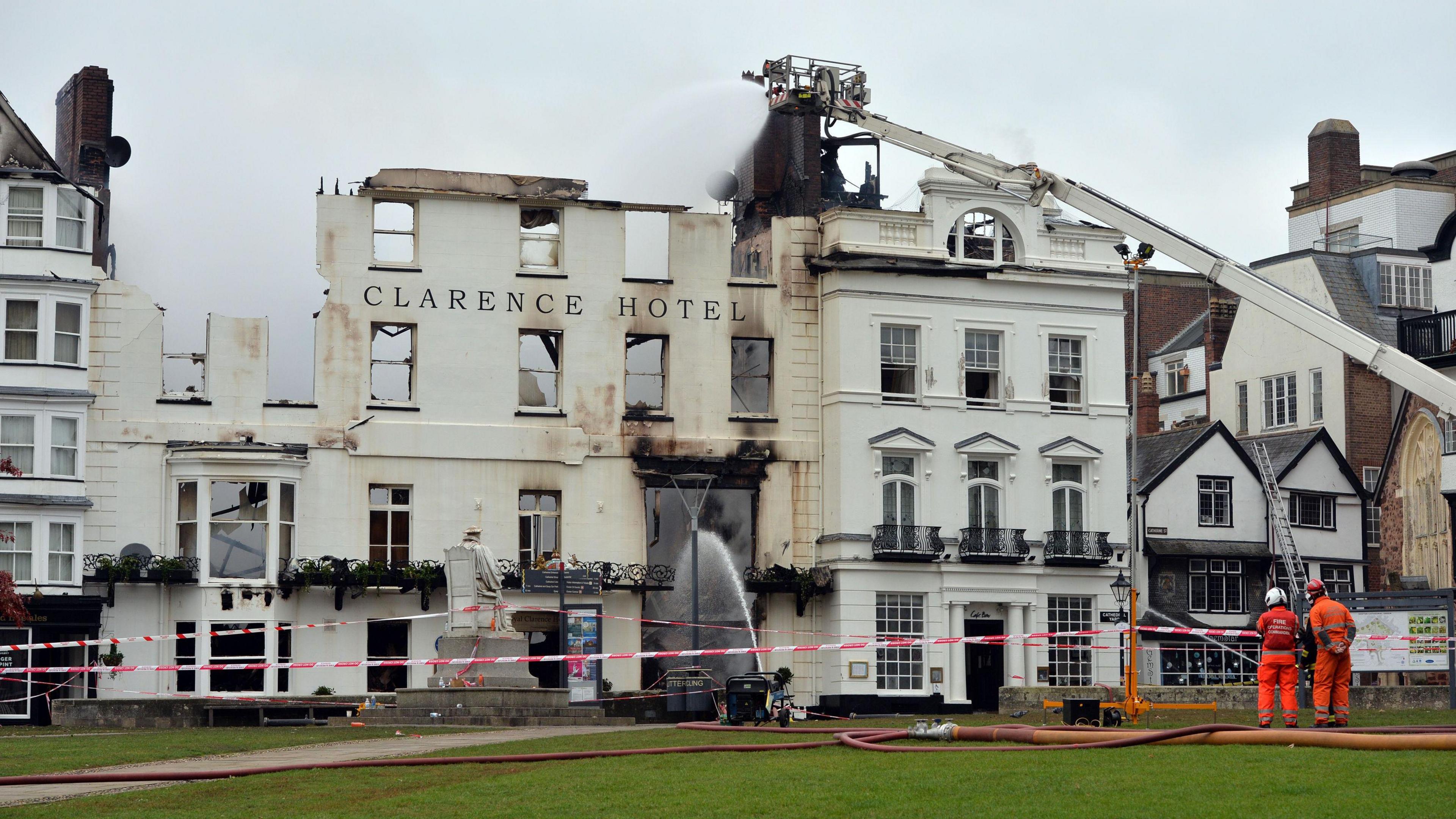 Firefighters in orange suits oversee water being sprayed from a crane onto the fire-damaged Royal Clarence Hotel. The four-storey historic building is partially burnt, with red and white tape cordoning off the area and a green lawn in the foreground.