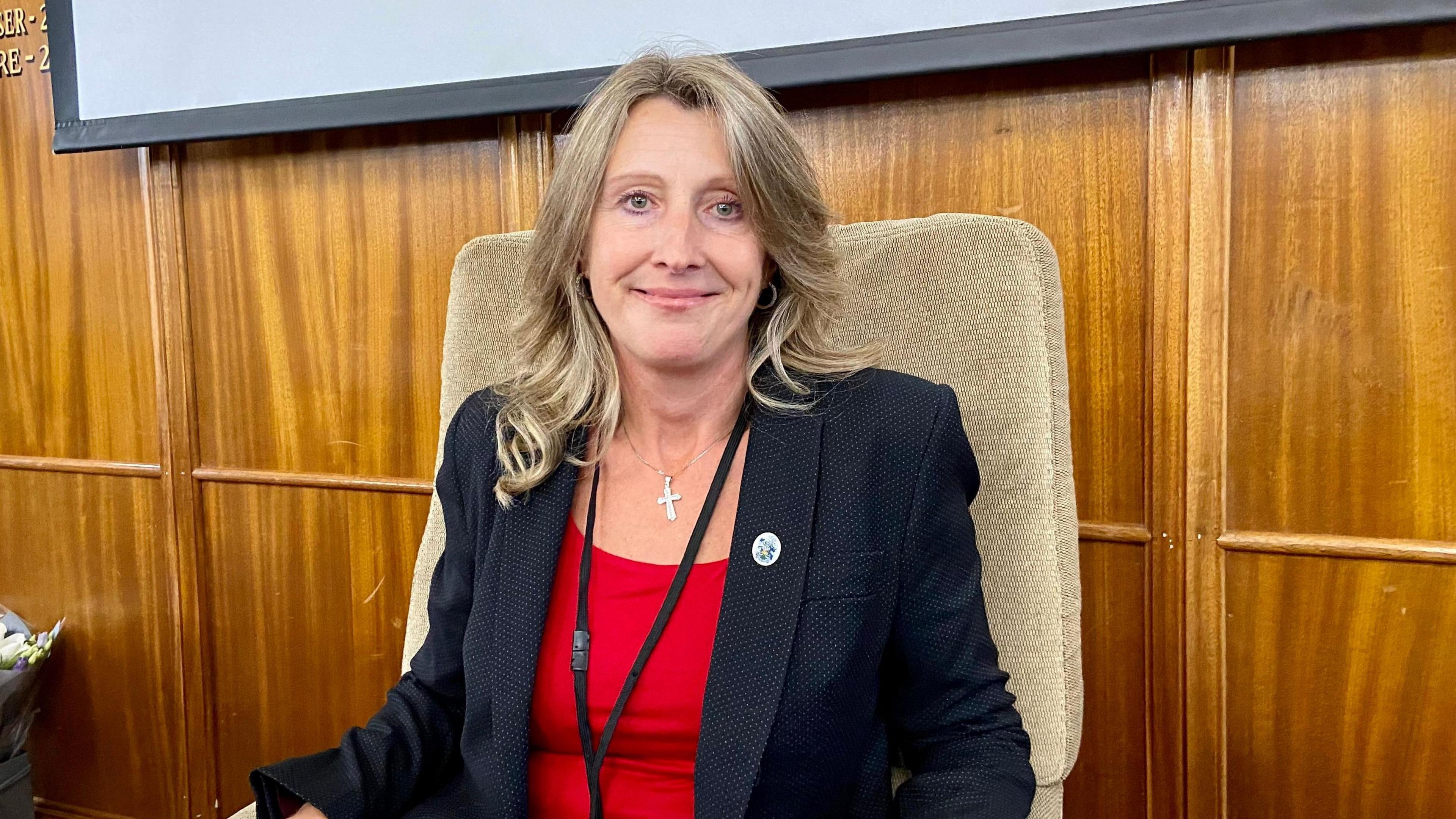 Joanne Sexton sits in a chair and looks directly at the camera, smiling. Behind her is a wood panelled wall and she is wearing a plain red top underneath a dark jacket