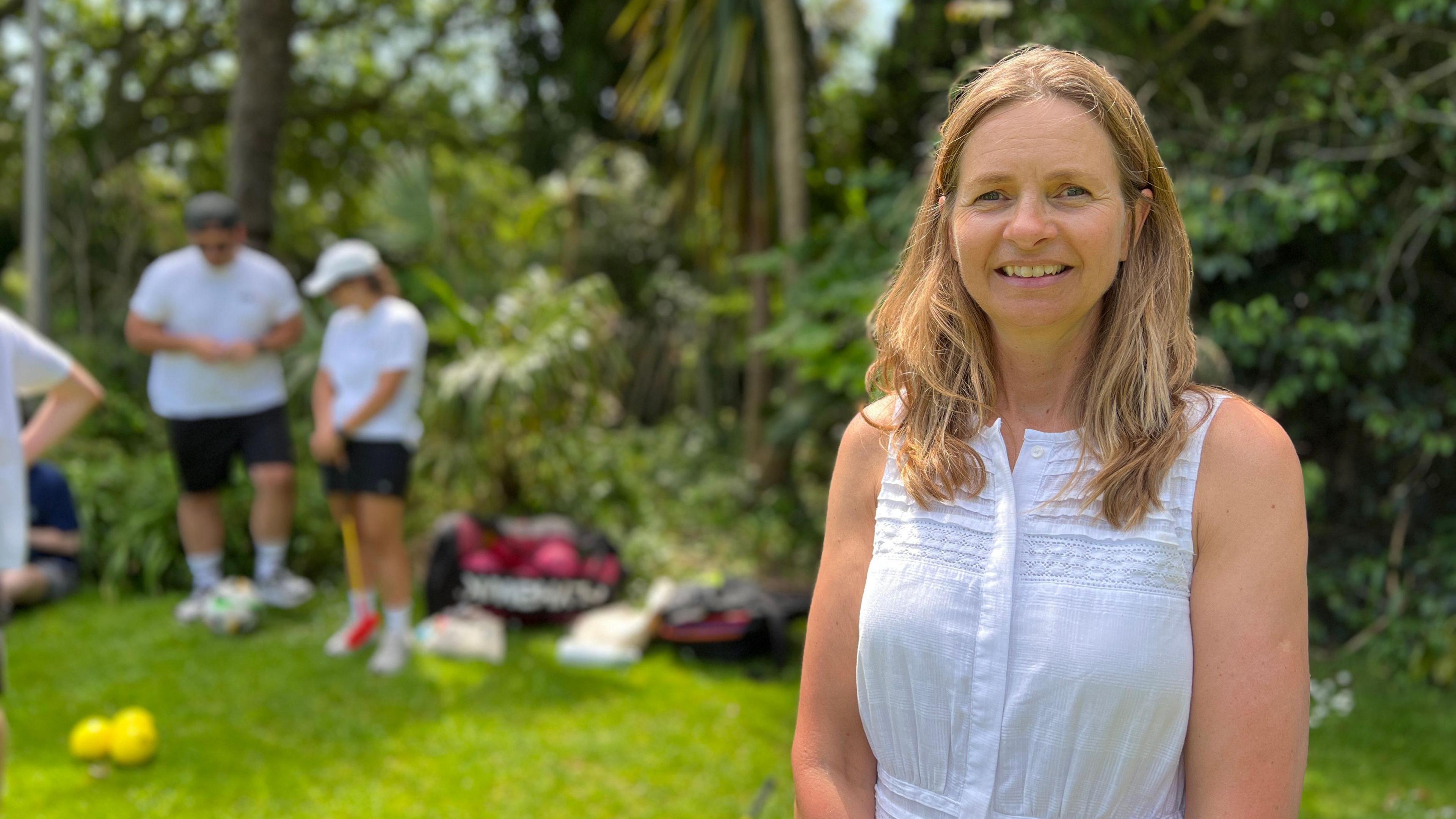 A woman, with shoulder length hair in a white top. She is looking at the camera smiling. She is is stood on the right. In the left in the background are other people and bags with sports balls in them. The grass is vibrant green and there are trees and bushes.