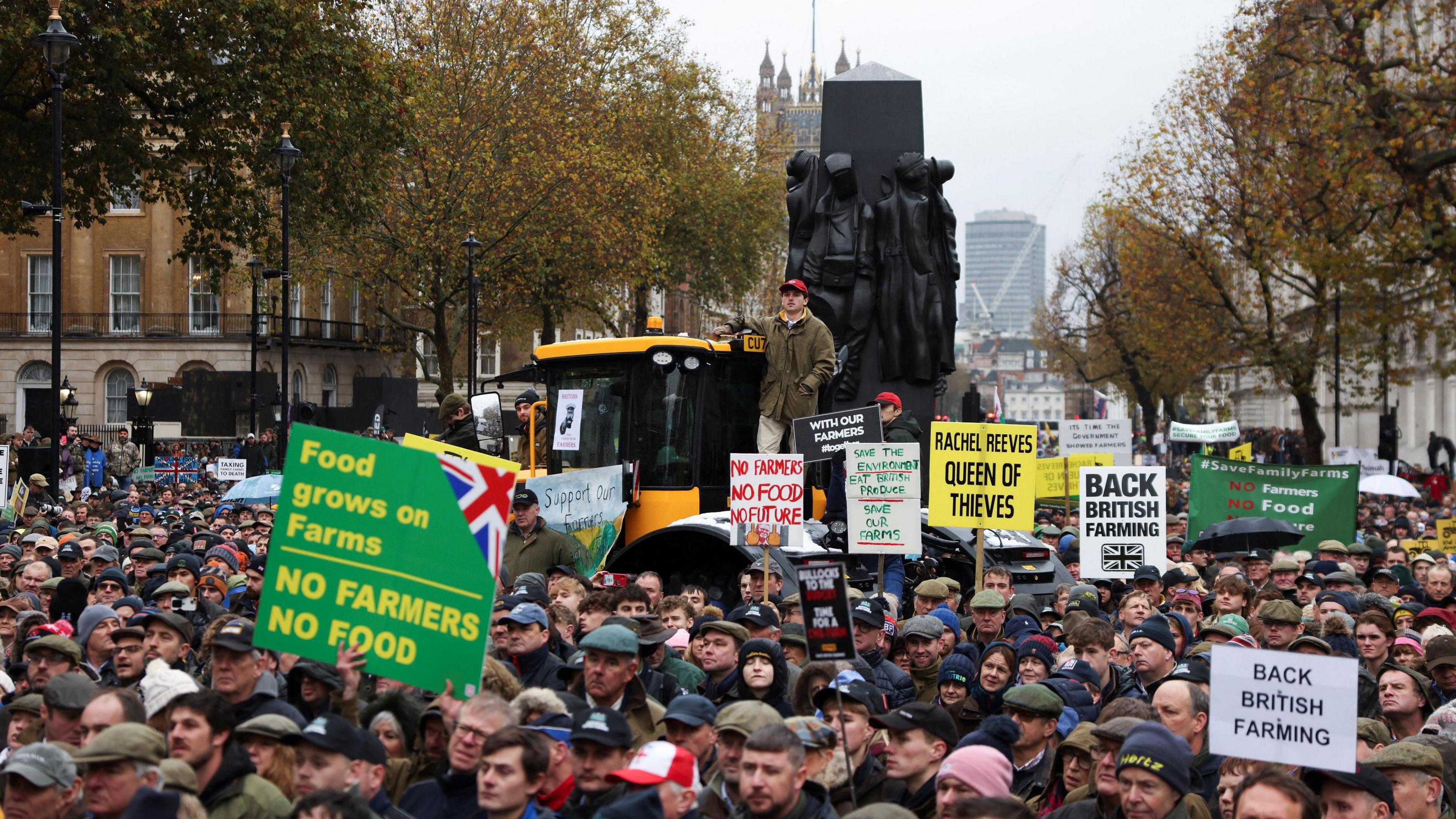 Farmers take part in a demonstration protesting against the Labour government's new agricultural policy. Some are holding up signs and a yellow tractor can be seen in the background.