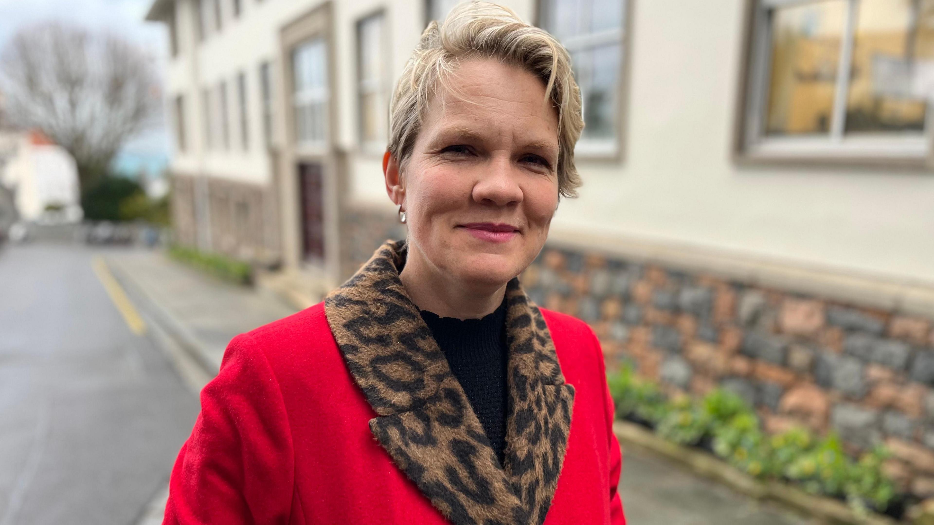 A lady with blonde haira and pink lipstick smiling towards the camera wearing a red coat with a leopard print lining and a black blouse. 