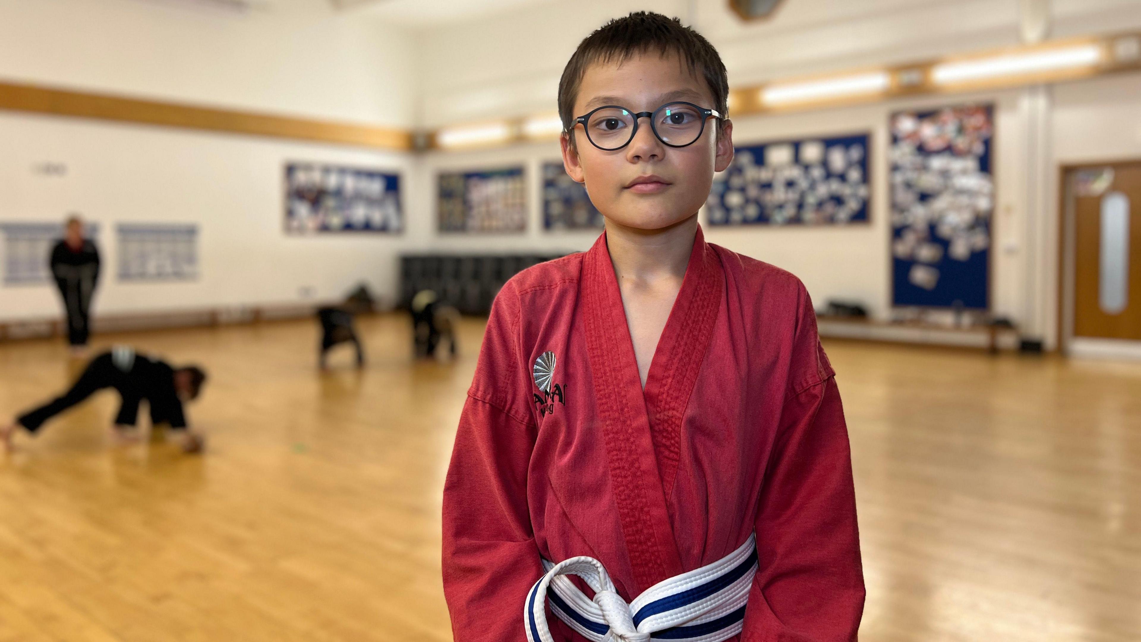 Jasper looks at the camera in his red gi with a Samurai Kickboxing logo on his chest. He wears glasses and has short dark hair and brown eyes. In the background children are doing press ups as a coach watches.