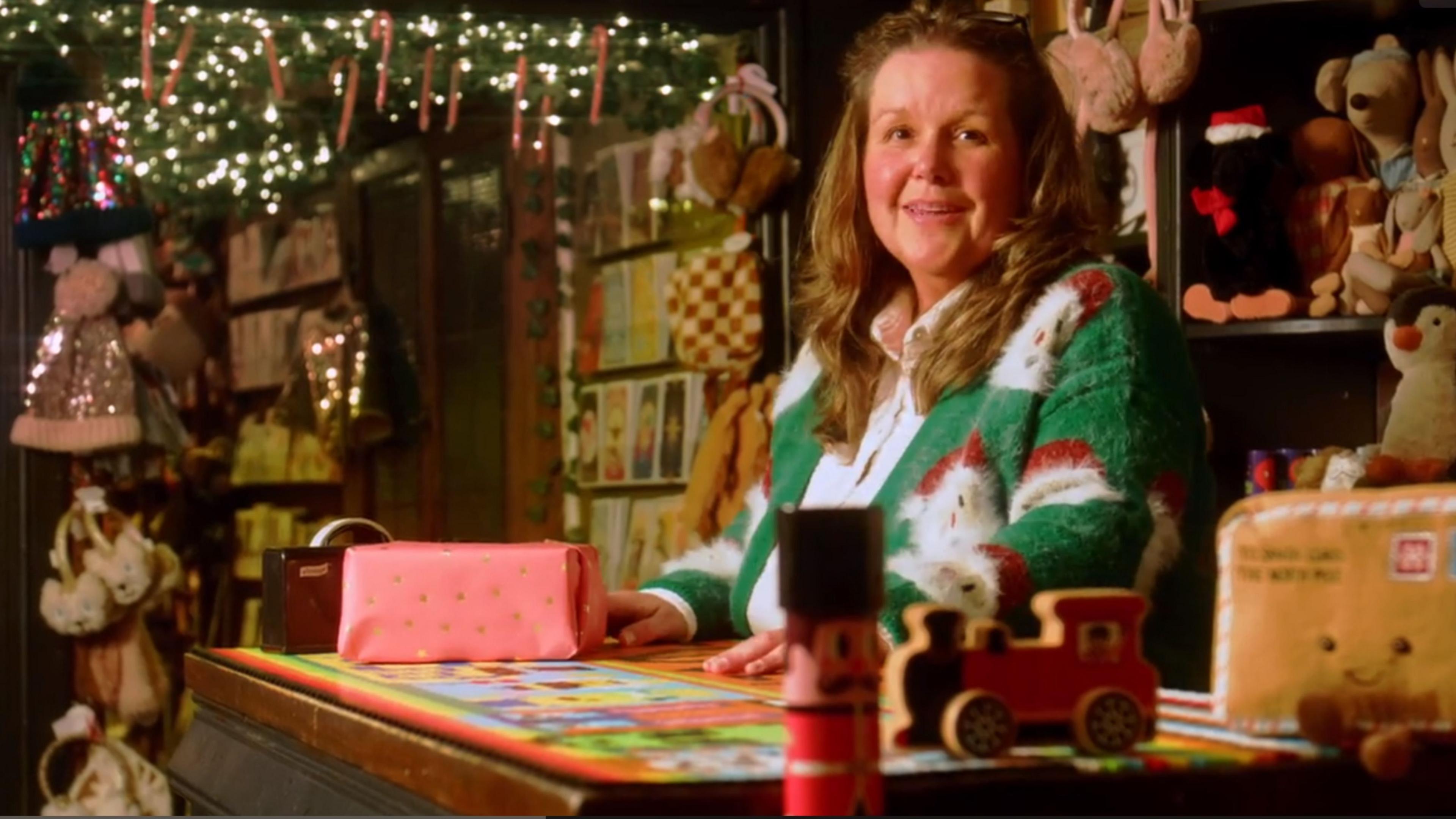 A woman with long pale hair stands behind a shop counter with a wrapped gift in front of her.
