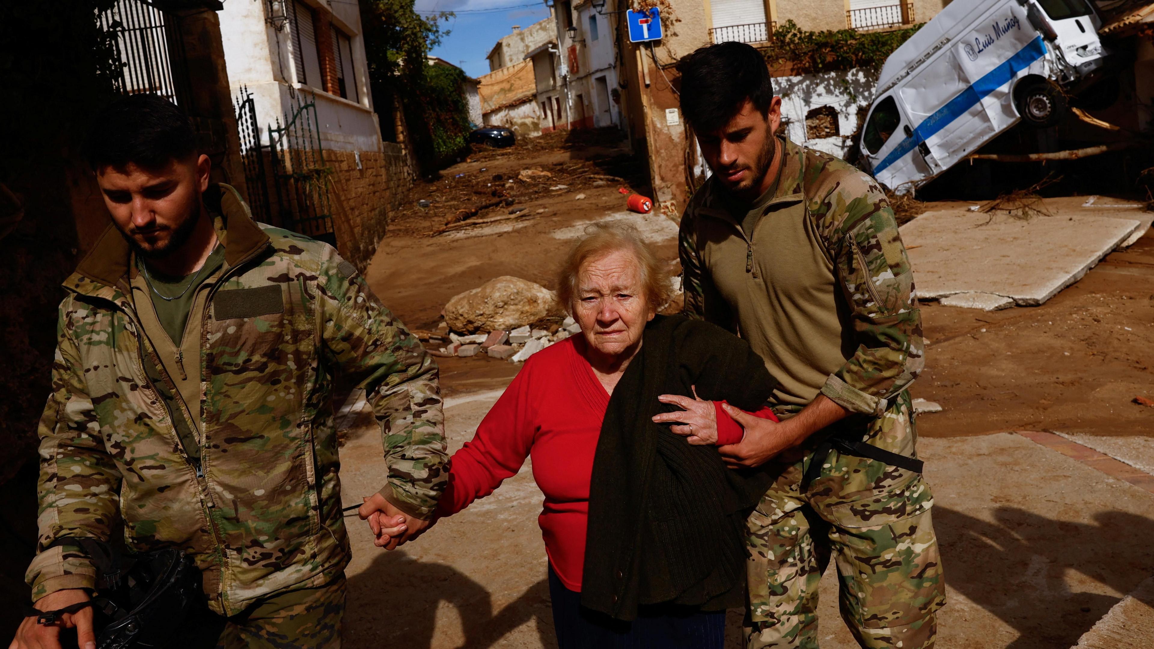 An elderly woman who looks upset is helped by two young men dressed in military fatigues. They hold her hands as she walks down a street covered in mud