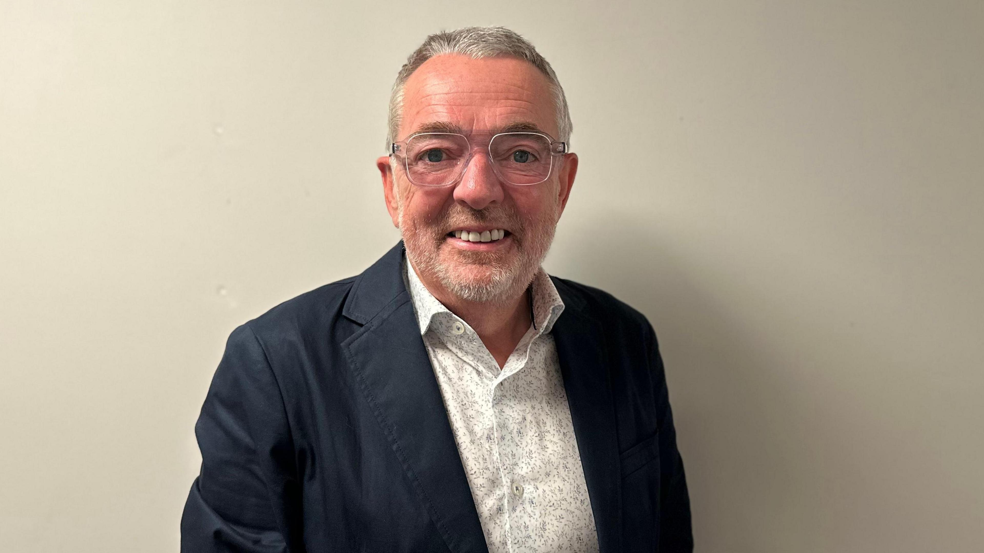 Prof Paul Seawright has short hair and stubble. He is wearing a white shirt with a floral pattern and a black blazer. He has silver-rimmed glasses and is smiling while standing in front of a plain wall.