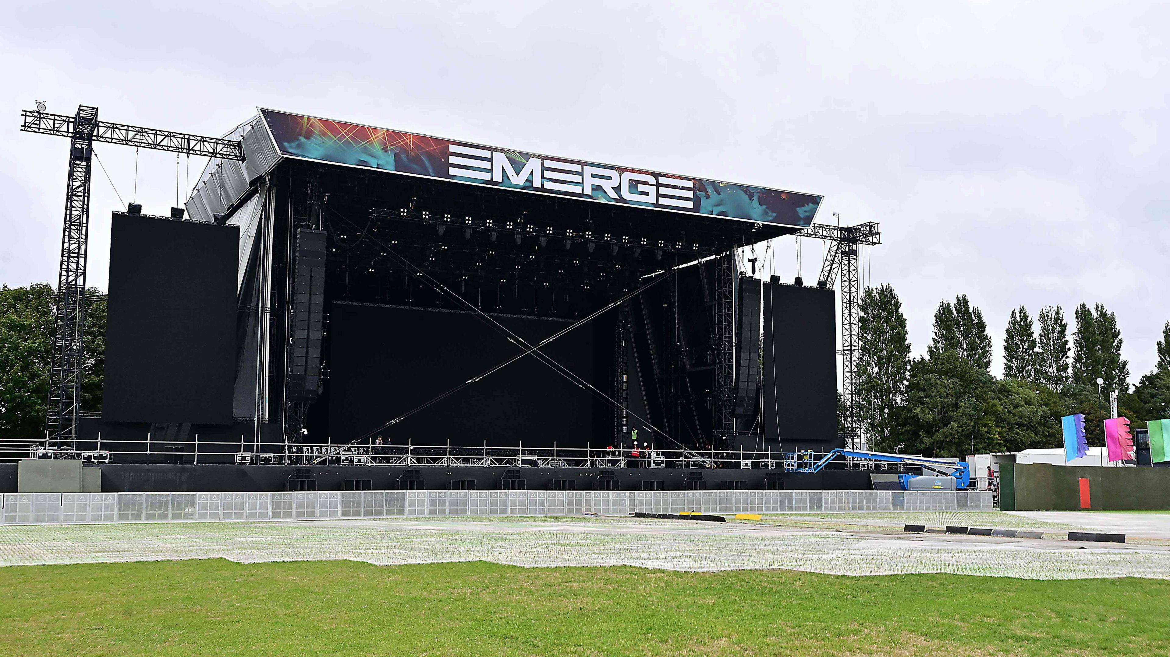 A large stage with a sign reading 'EMERGE' above it. There are a number of trees to the right of the stage, and a tree to the left of the stage. The sky is overcast.