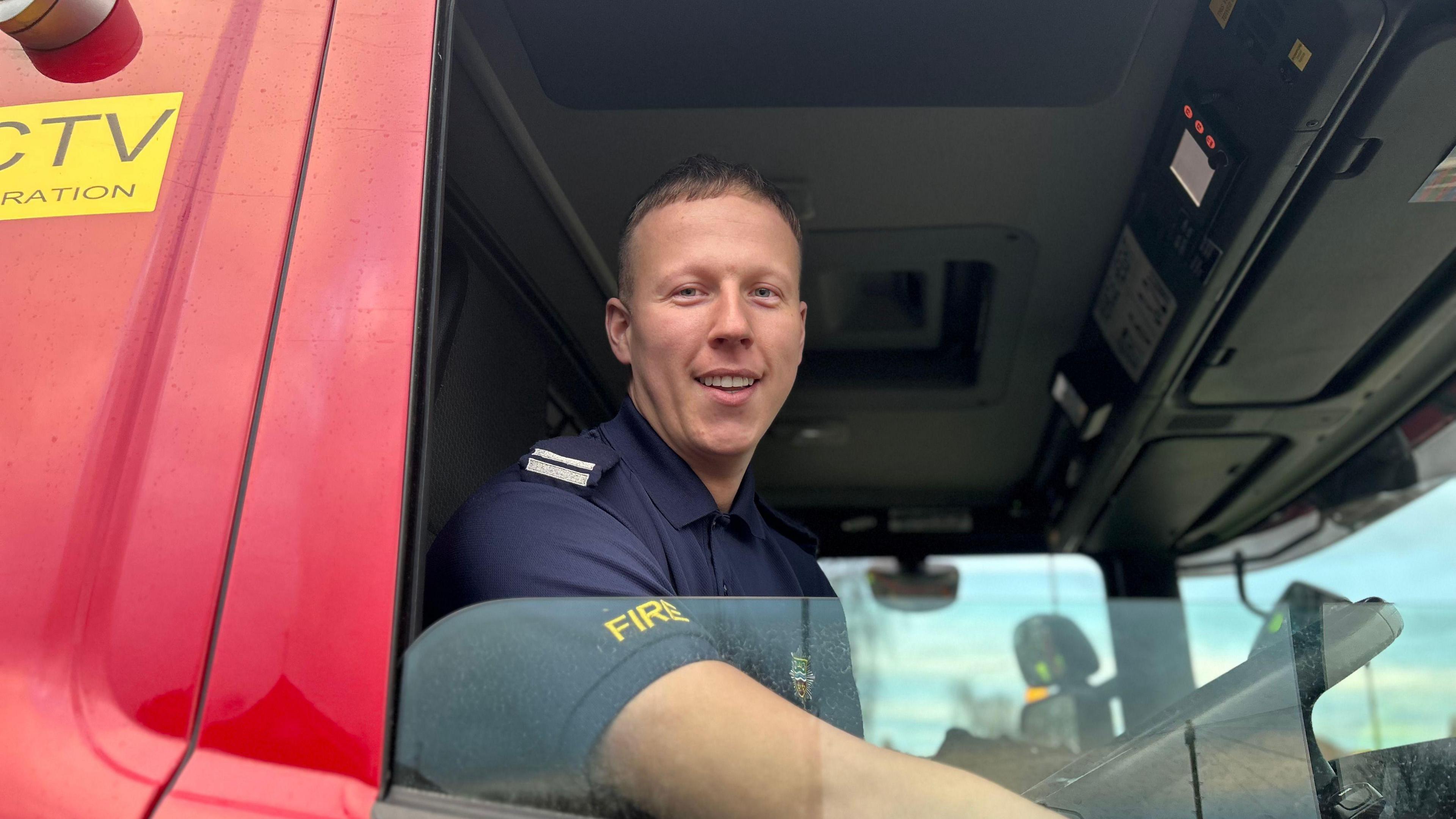 A firefighter sitting in the driver's seat of a fire engine, looking through the window, which is down, and smiling