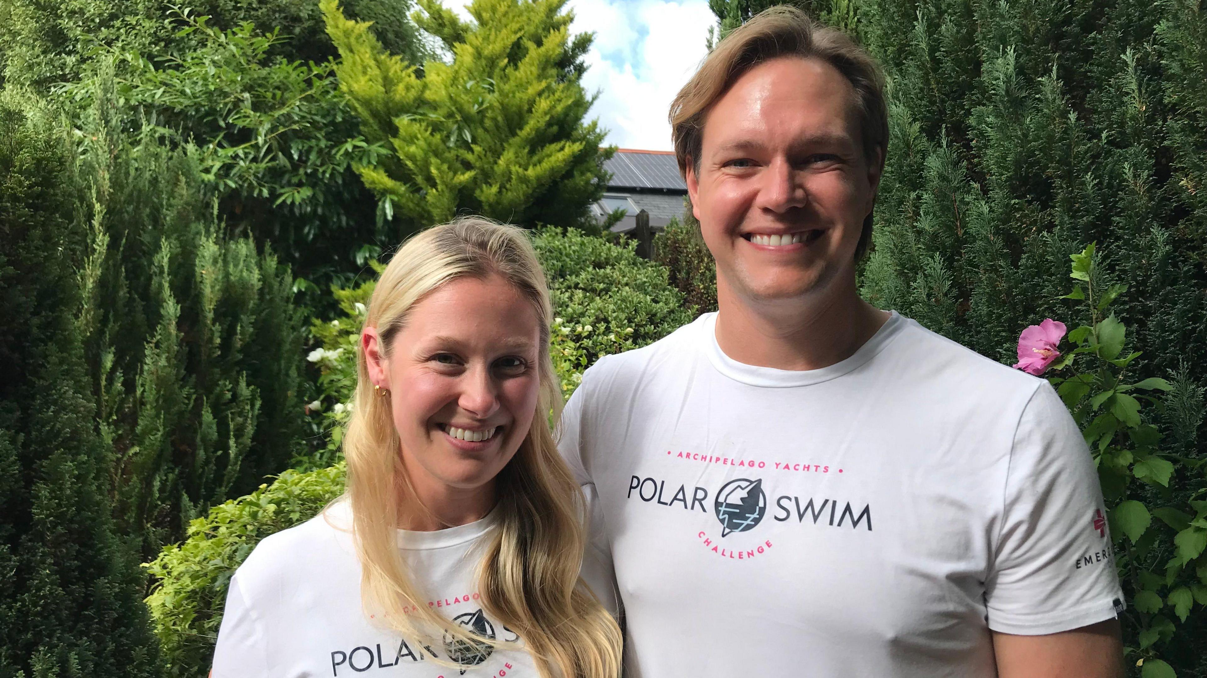 A woman, on the left, with mid length blonde hair and a man with short hair, both smiling at the camera. They are both wearing white t-shirts that say "Polar Swim" on them. Behind them are green bushes. 