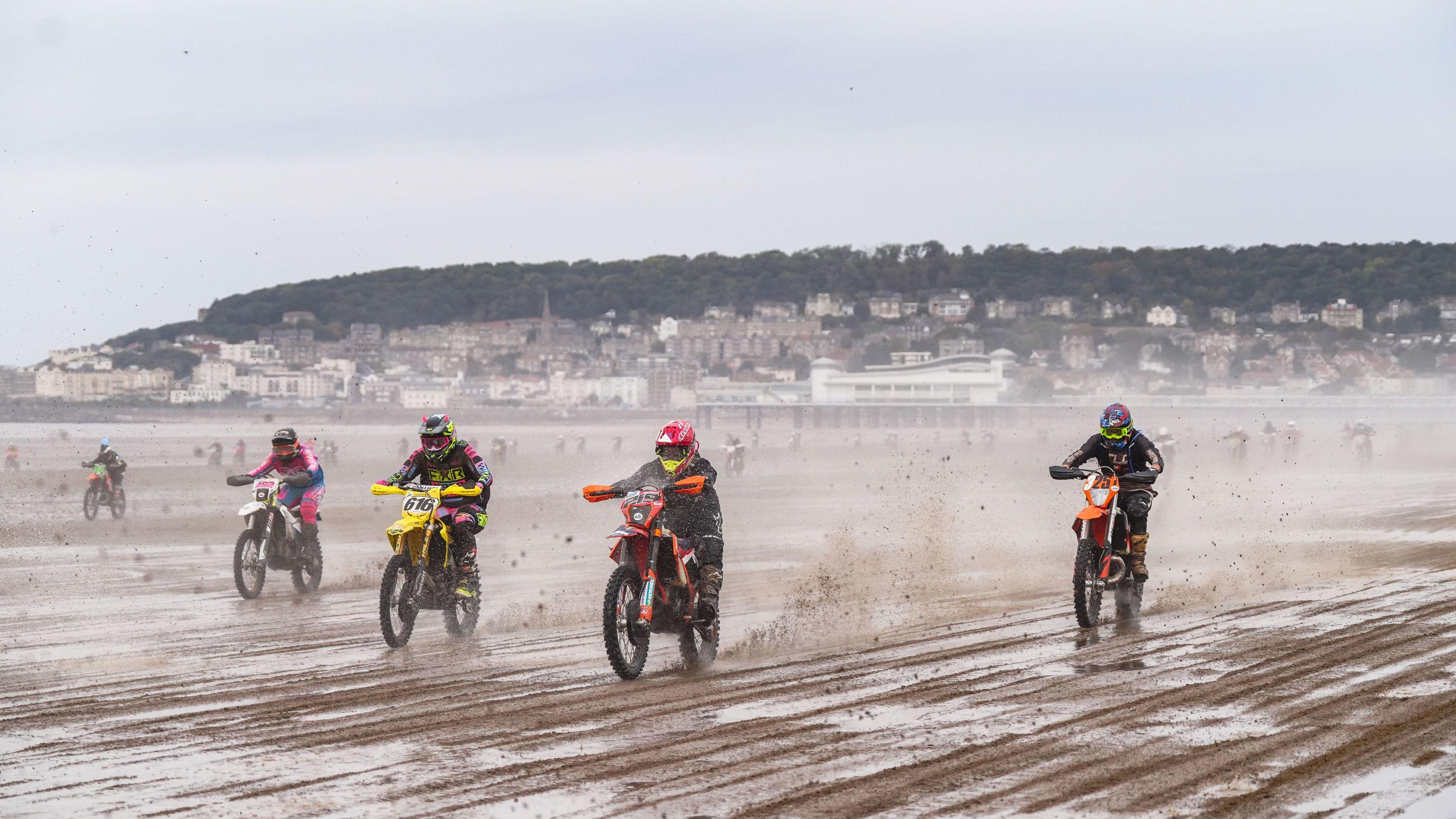 Several motorbike riders race across wet sand during the Weston Beach Races at Weston Super Mare. The town's pier is visible in the background