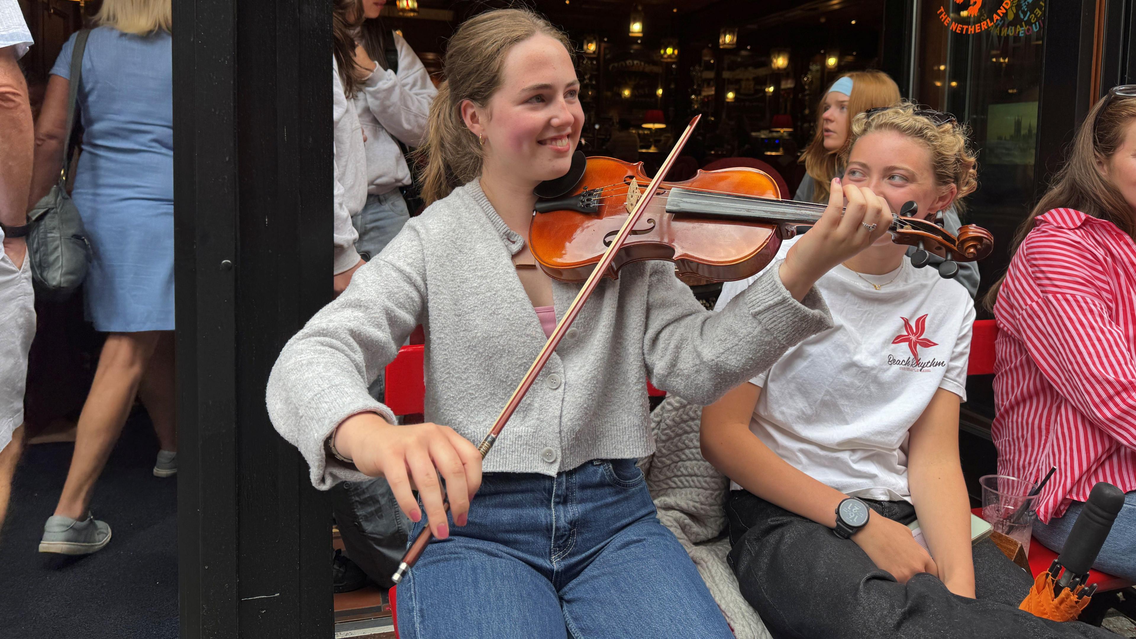 A girl with brown hair playing the violin, she has people sitting beside her watching