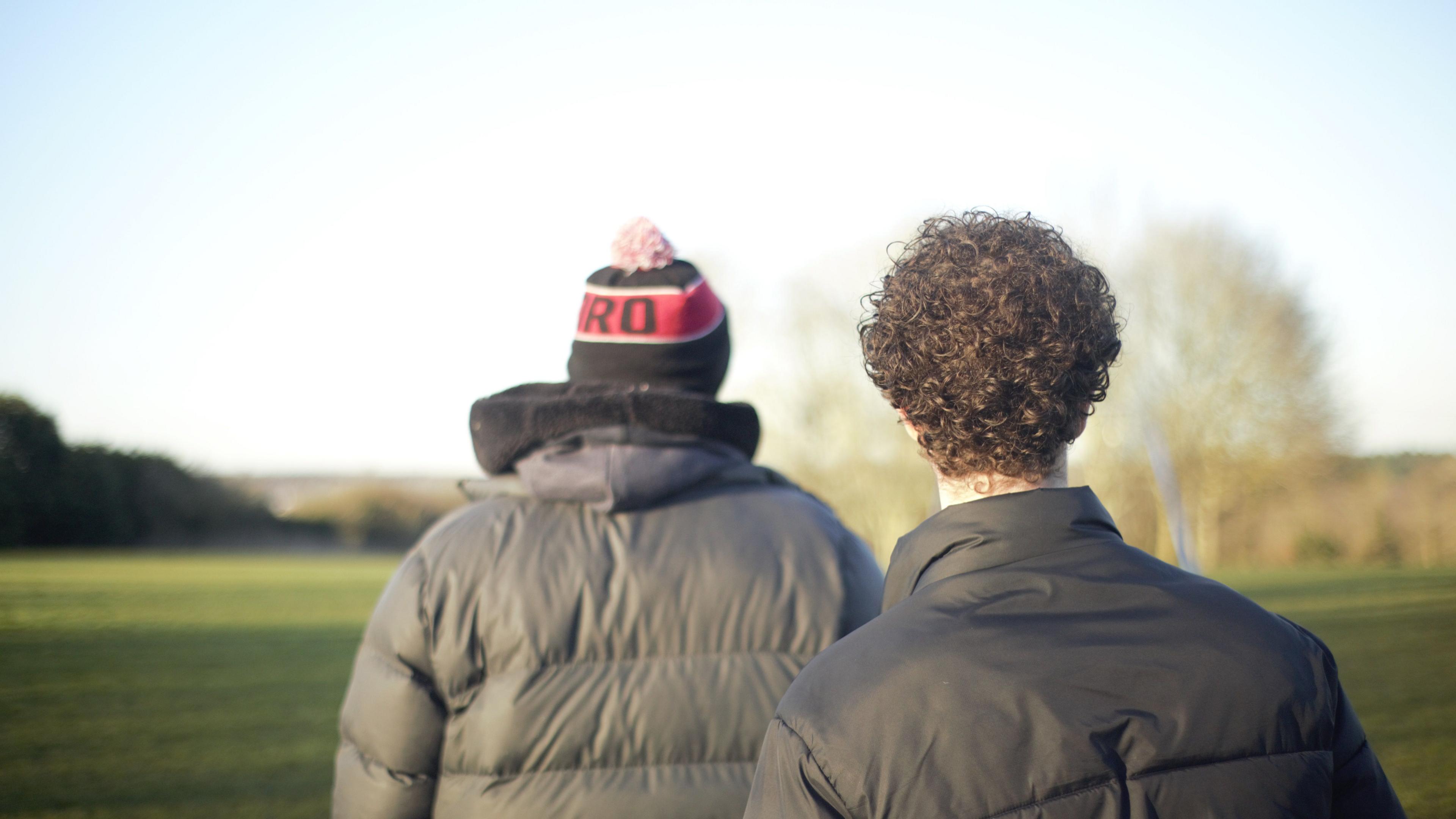The back of John and Oscar. John is wearing a hat, with a bobble, on and a dark coat, Oscar, has short curly hair and is wearing a black coat. They are standing in a field, with grass and trees in the distance. 