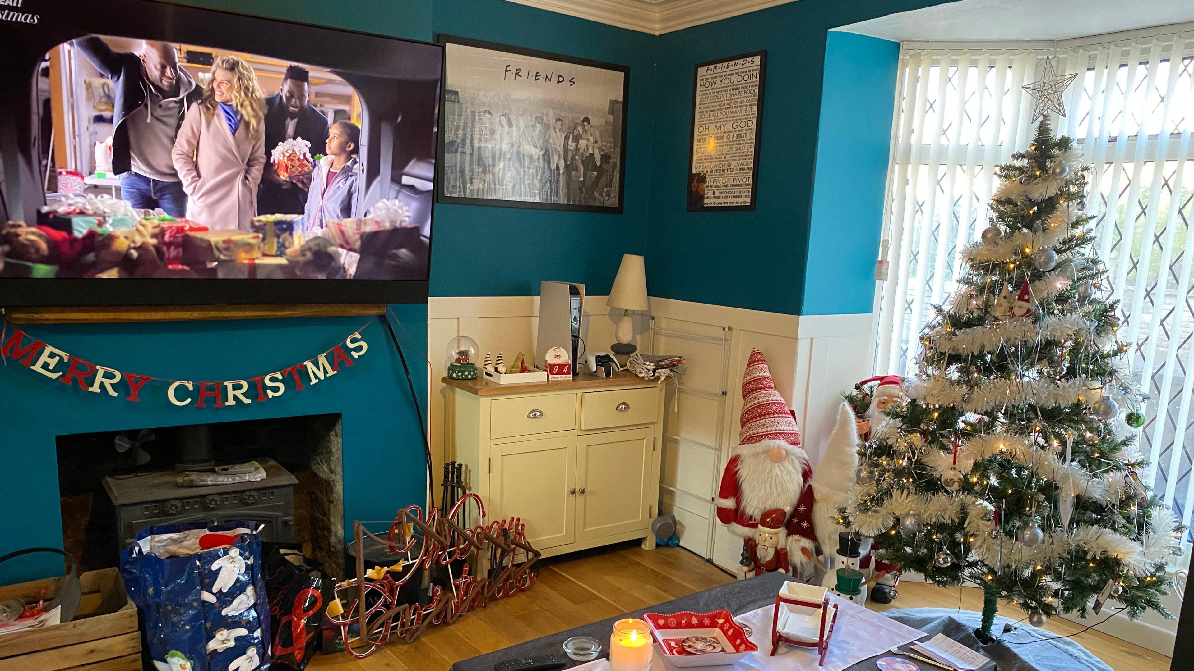 A front room is decorated for Christmas. A tree is in the bay window, with some Santa figures next to it. A Merry Christmas garland has been hung under the TV.