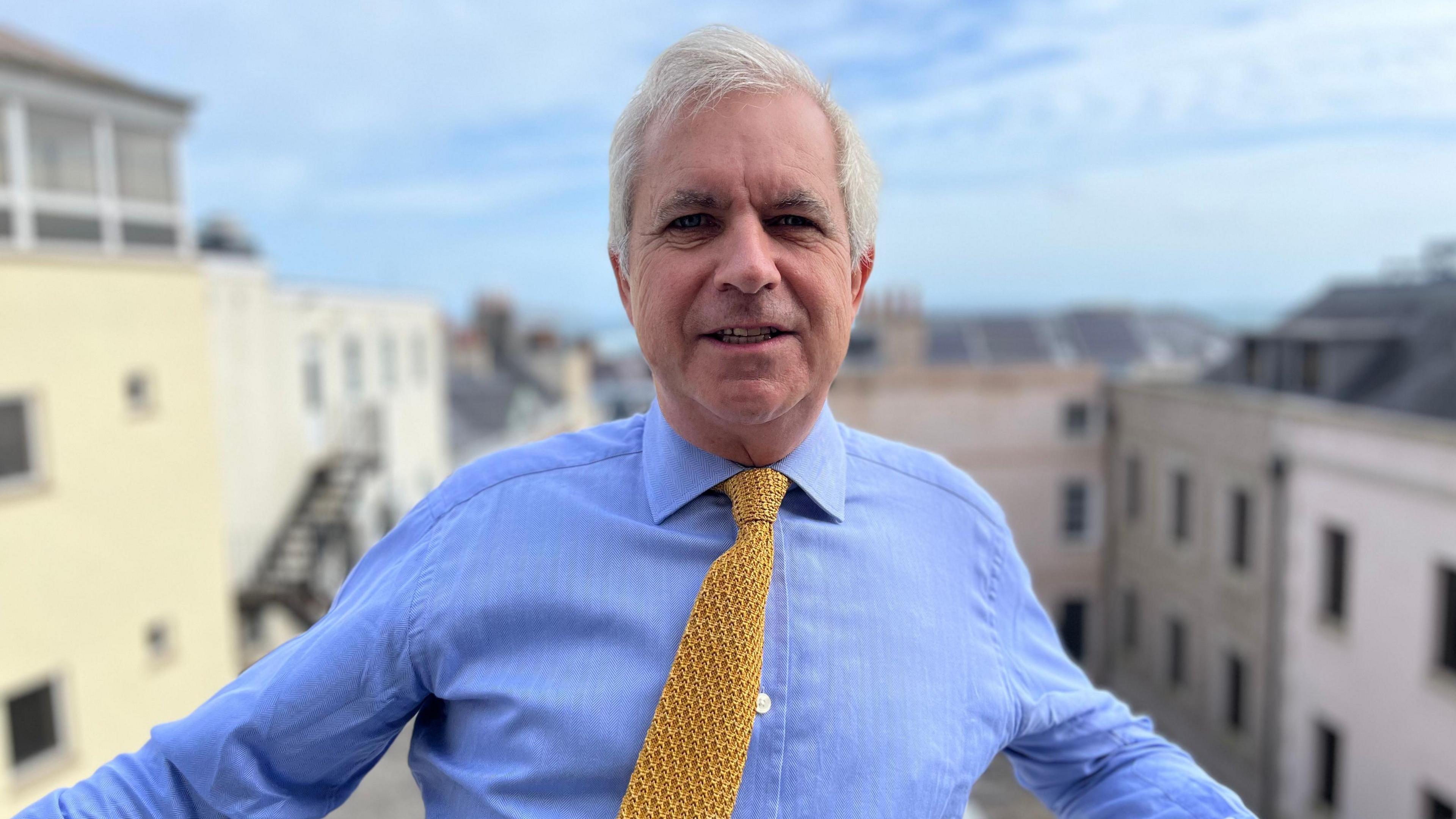 Christopher Beaumont wearing a blue shirt and yellow tie. He is standing outdoors. Buildings are in the background.