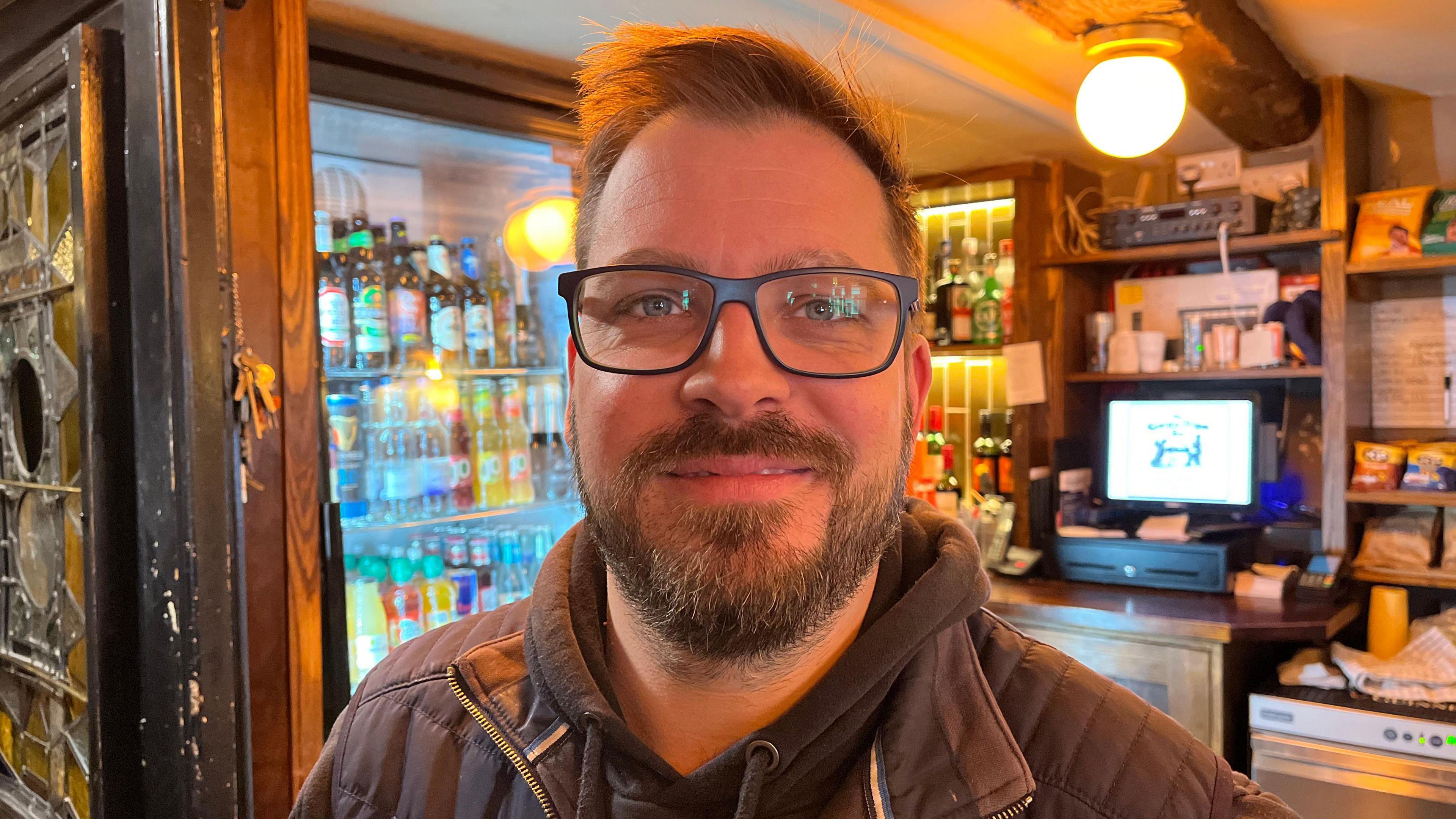 A man with spikey brown hair and it wearing glasses. He is smiling and has a beard. He wears a dark jacket with a black hoodie underneath. He is standing in front of a bar inside a pub